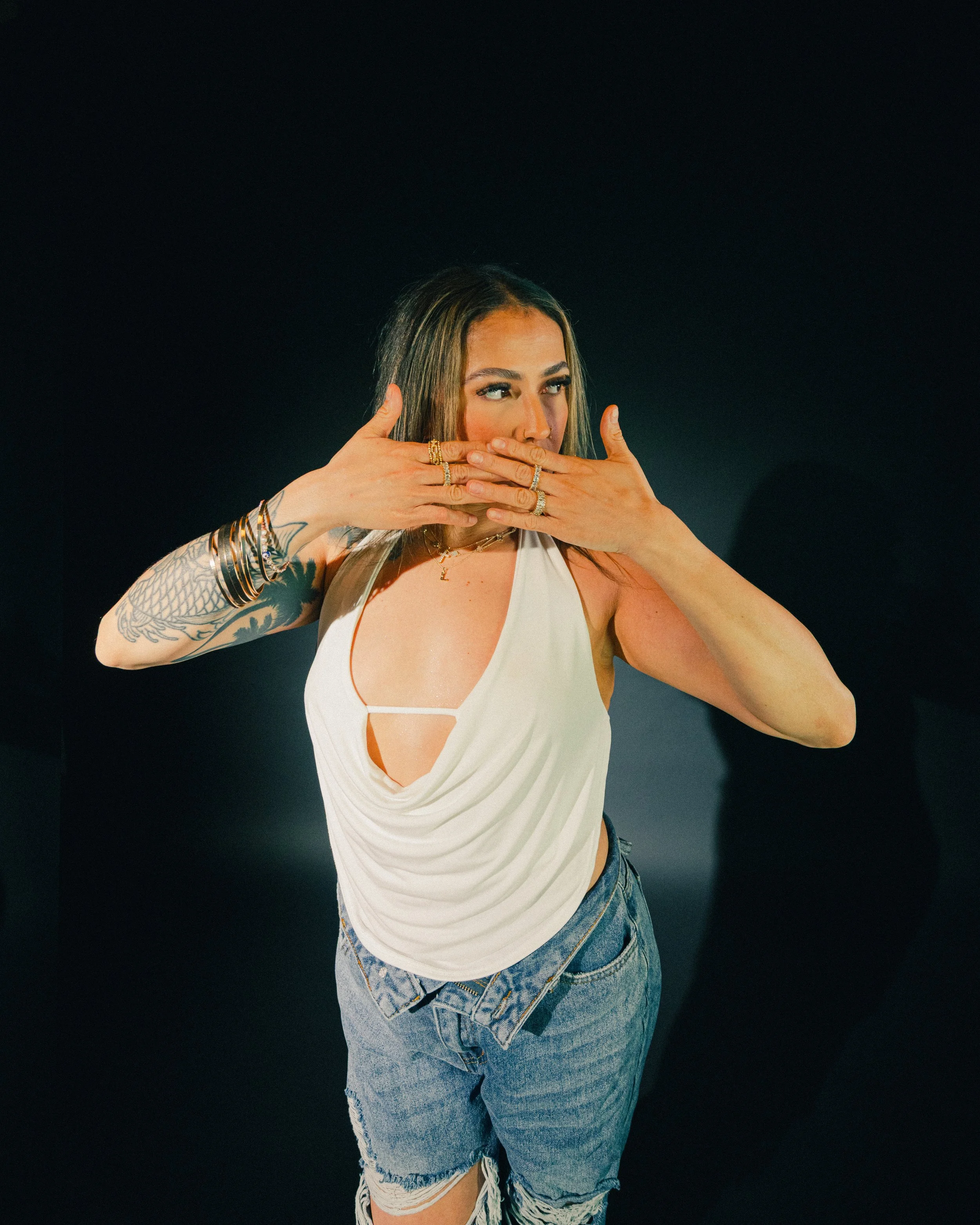 Young woman with long hair, tattooed arms, and jewelry, wearing a white sleeveless top and ripped jeans, posing against a black background.