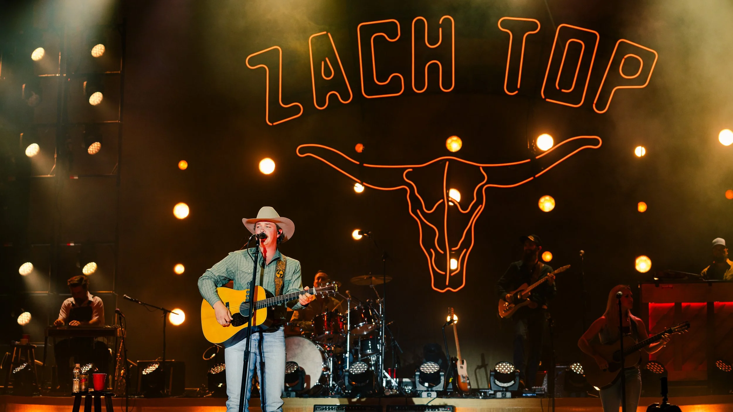 A musician singing and playing guitar on stage during a live concert with a neon sign above him that reads 'TACHA TOP' and a longhorn steer skull outline. The stage is lit with multiple round yellow and orange lights, and band members are visible in 