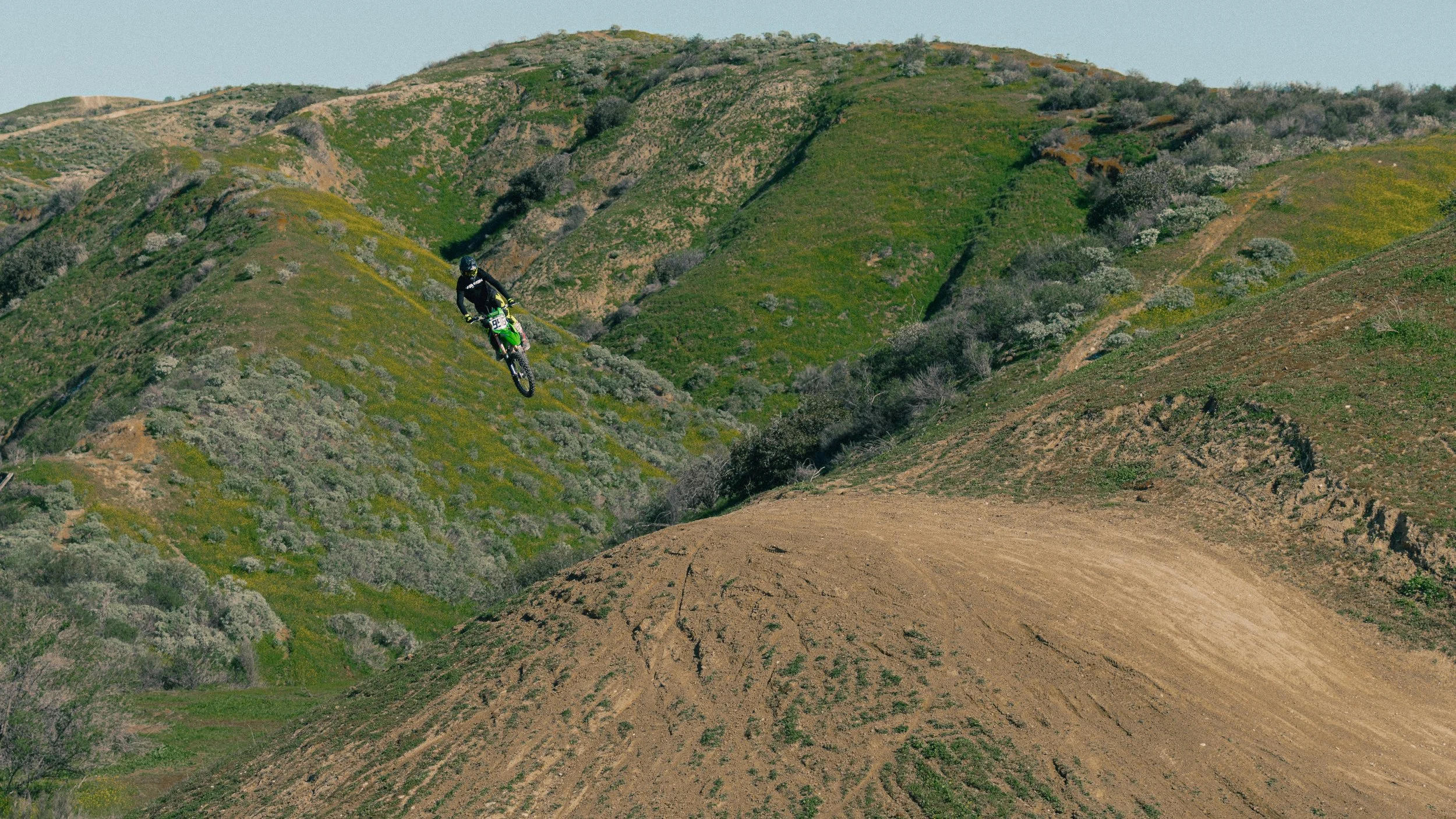 A mountain biker wearing a helmet and black gear riding down a dirt trail on a green hillside with sparse bushes and patches of yellow and purple wildflowers.