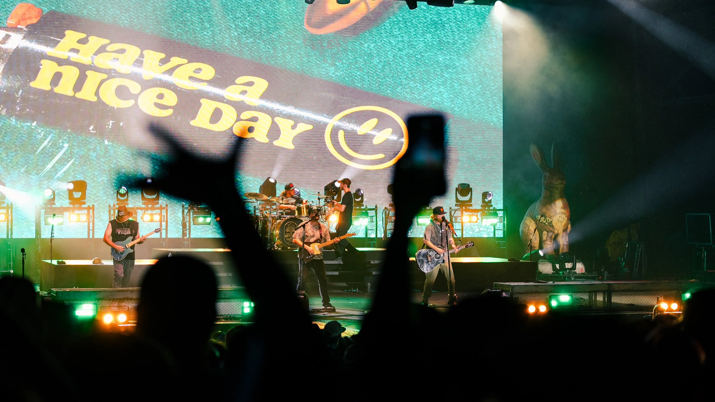 A live music concert with multiple band members playing guitars and drums on stage. Audience members are raising their hands in the foreground, with a large screen behind displaying the words "Have a Nice Day" along with a smiley face. Bright stage l