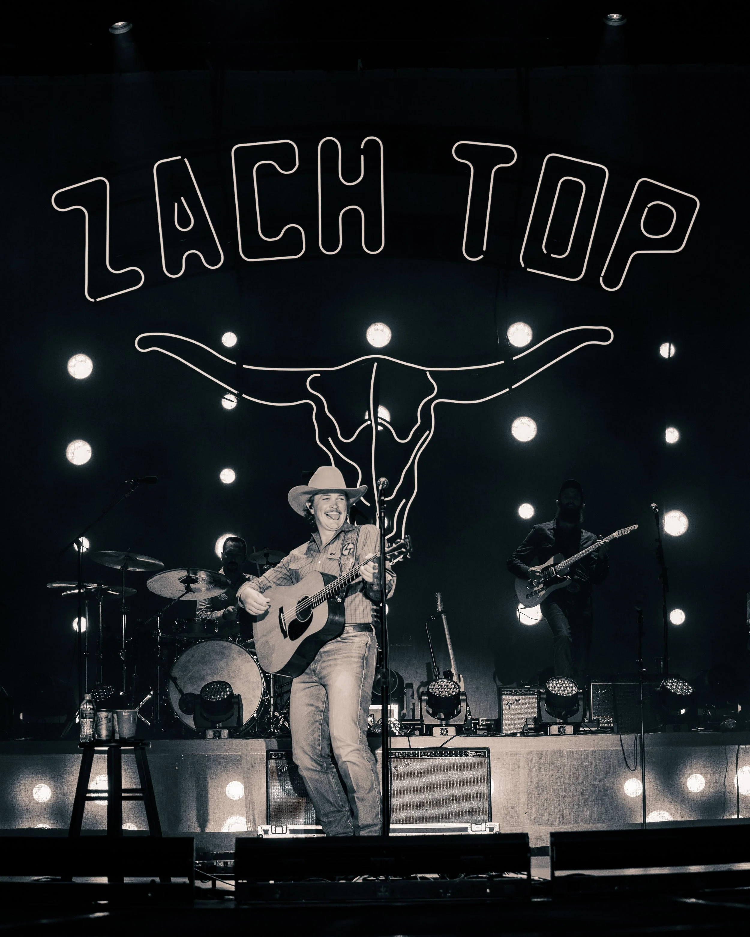 Musician performing on stage with a guitar, accompanied by a band, in front of a backdrop with a neon sign reading 'Lach Top' and a bull skull graphic.