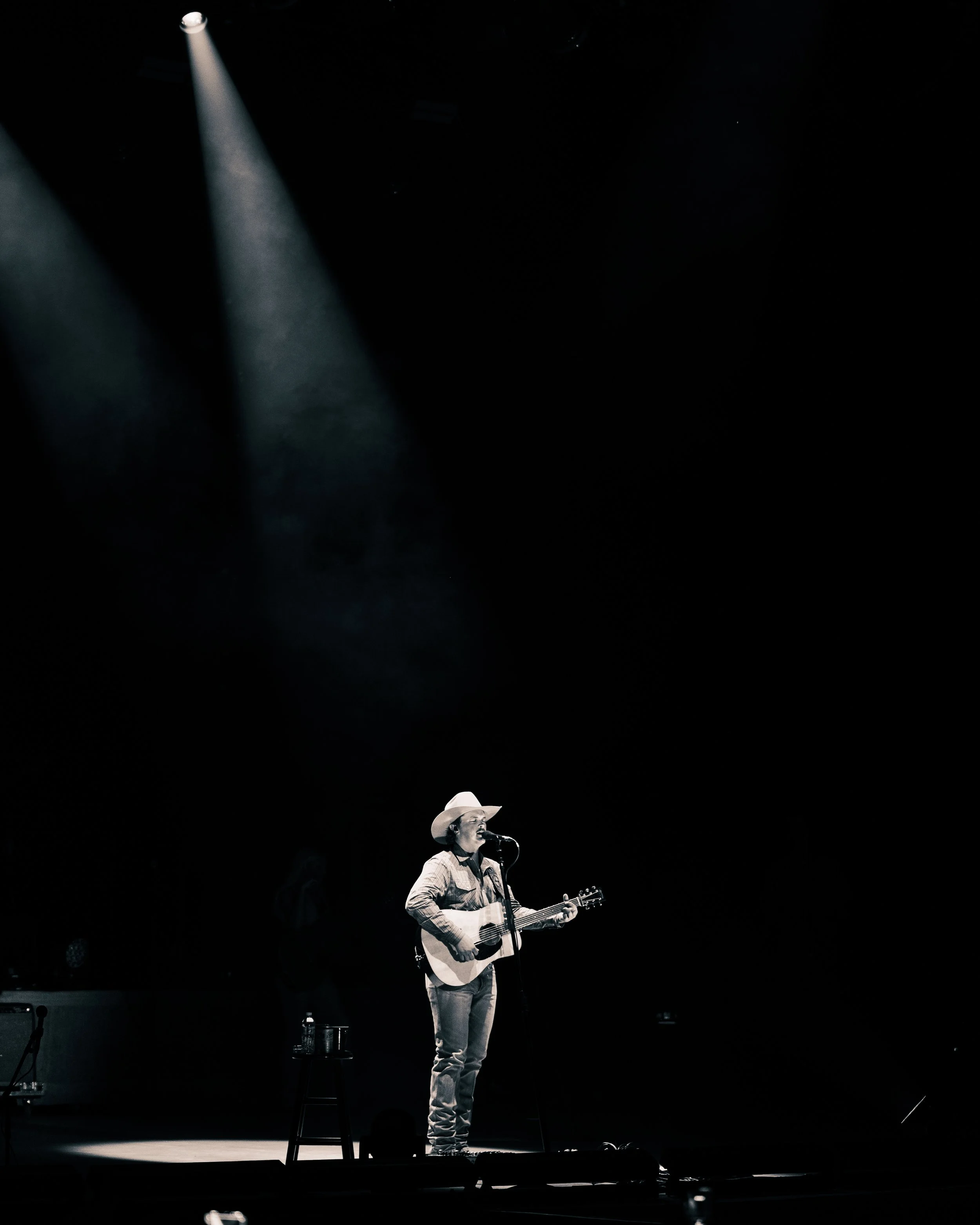 A man in a cowboy hat singing into a microphone while playing an acoustic guitar on a dimly lit stage.