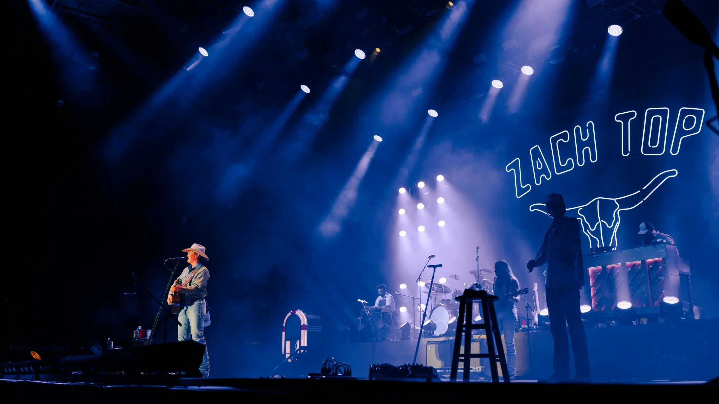 A stage with musicians performing, illuminated by blue and purple stage lights. A sign reading 'Lach Top' is displayed in the background. Some musicians are playing instruments, including a guitarist in the foreground wearing a cowboy hat.