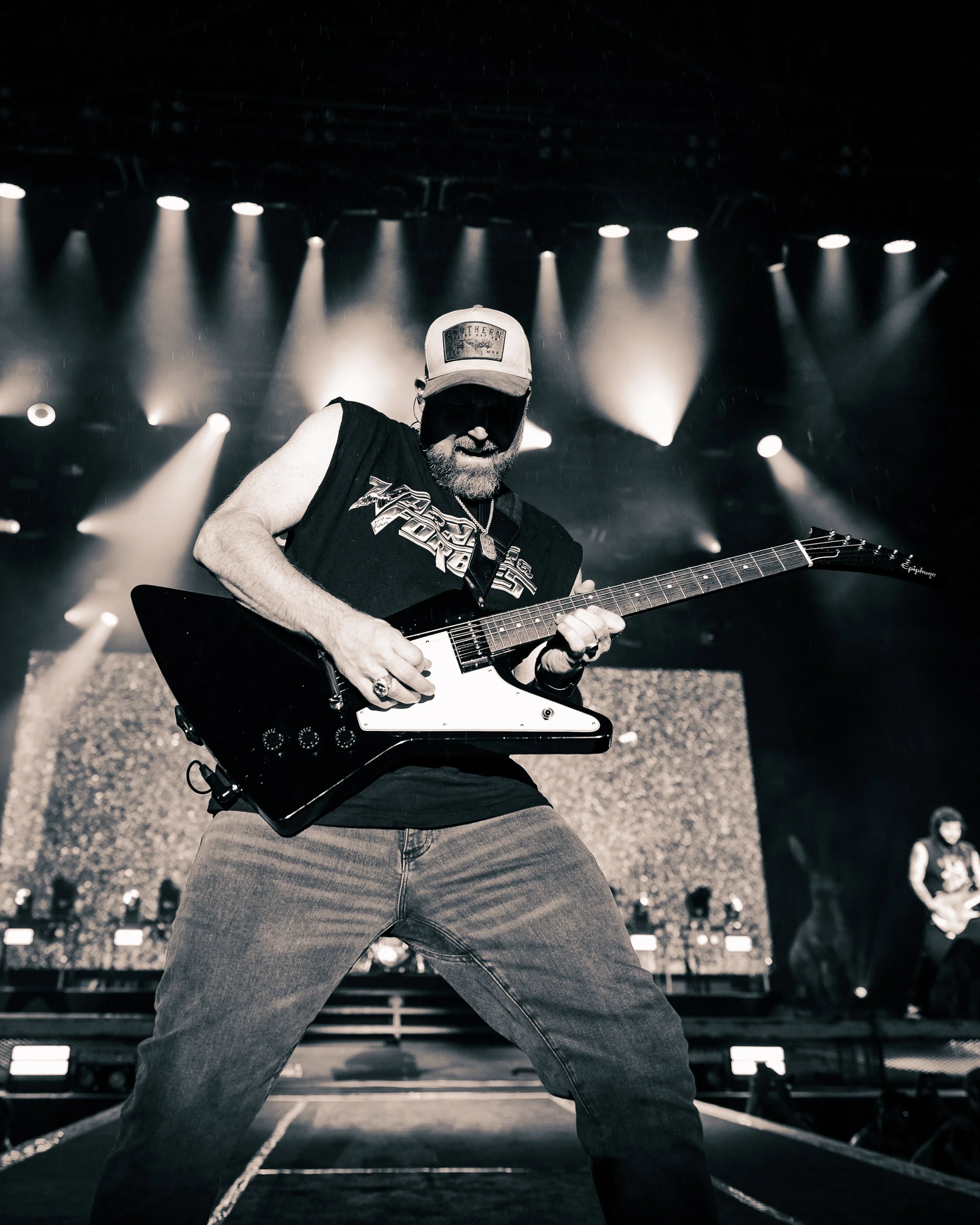 Black and white photo of a man playing an electric guitar on stage under bright spotlights, wearing a baseball cap, sunglasses, a sleeveless shirt, and jeans.