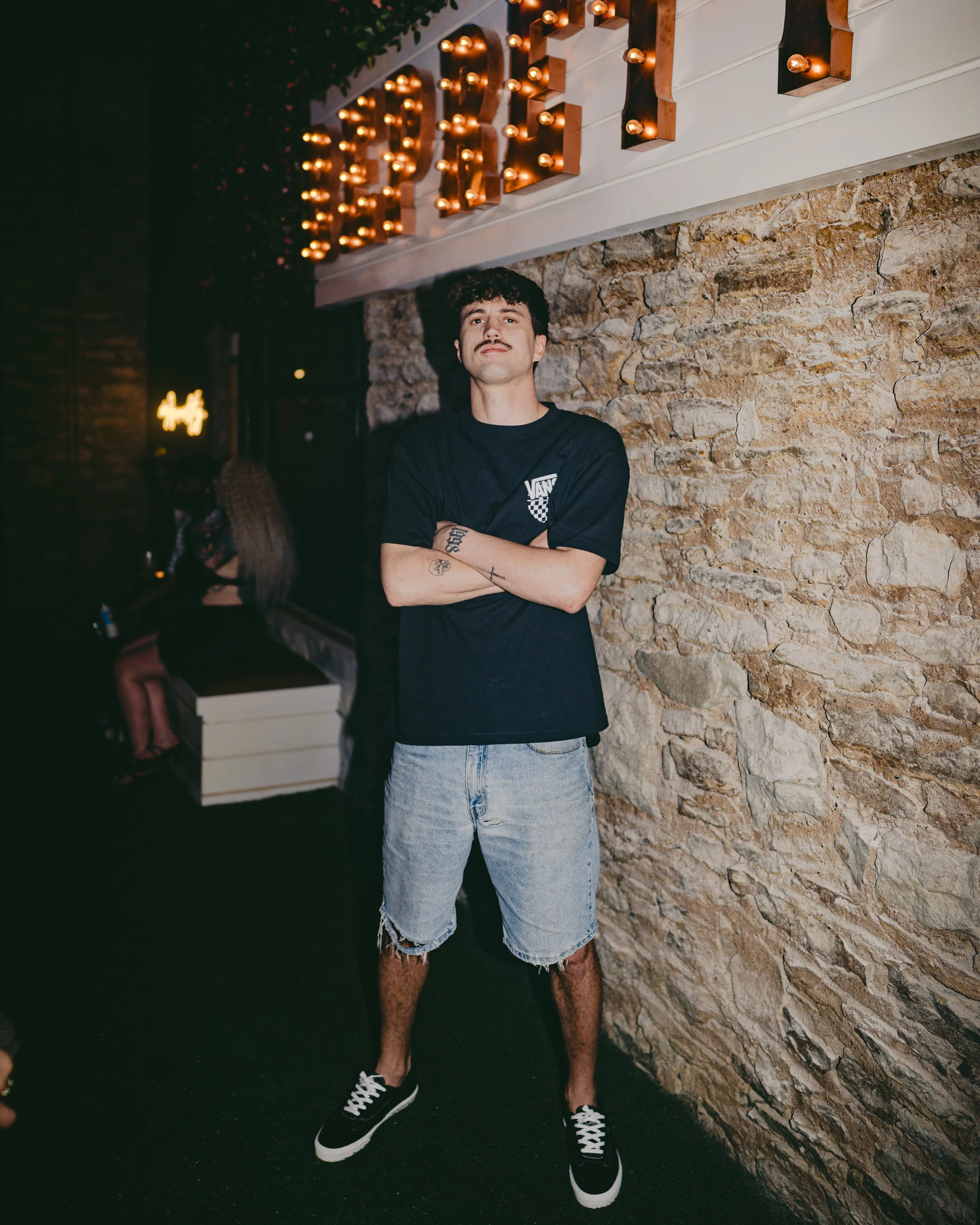 A man standing with crossed arms in front of a stone wall illuminated by a lit marquee sign, wearing a black T-shirt, ripped denim shorts, and black sneakers.