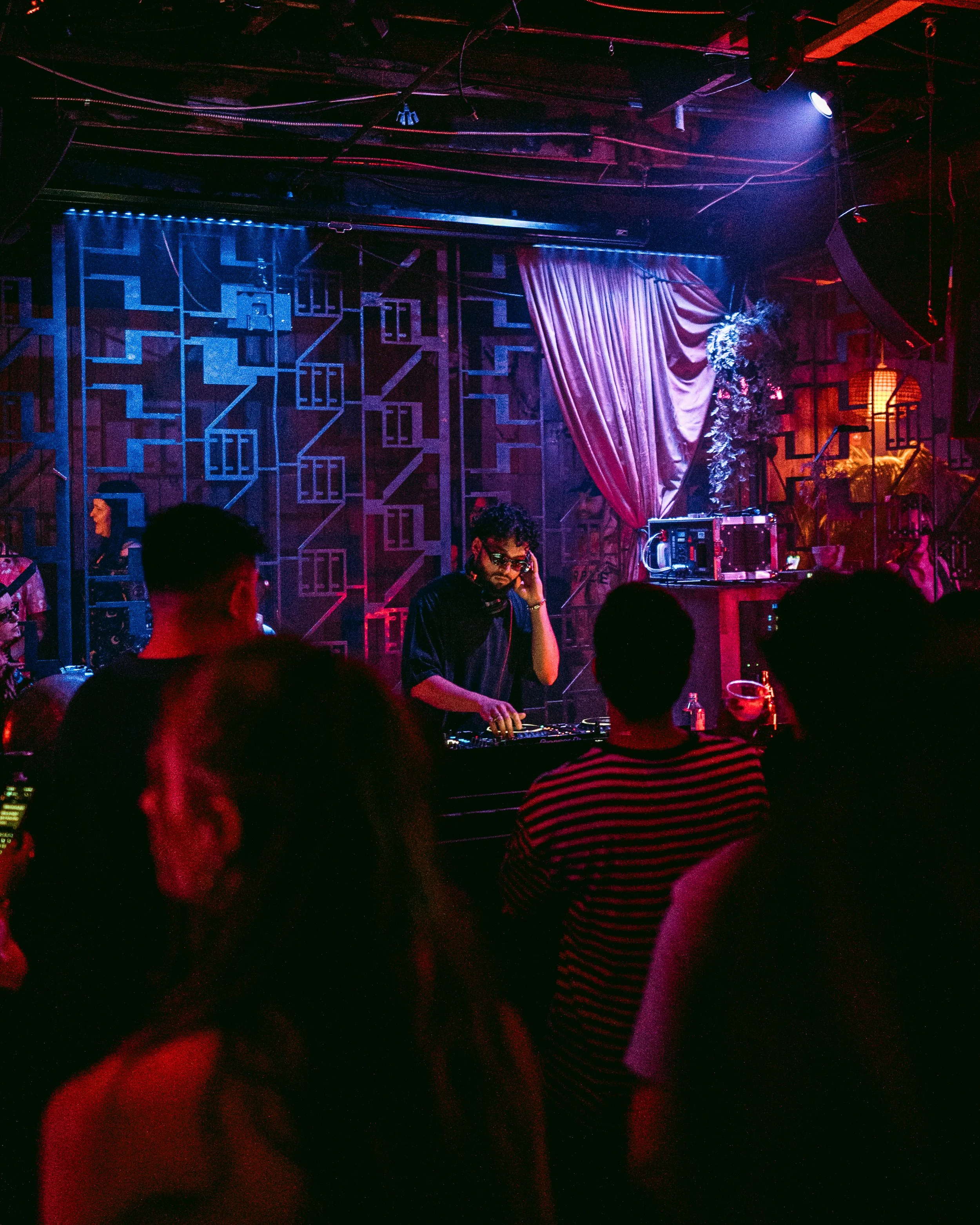 A DJ performing at a nightclub with a crowd of people dancing and socializing, dimly lit with colorful neon and purple lighting, decorated with elaborate drapes and geometric wall art.