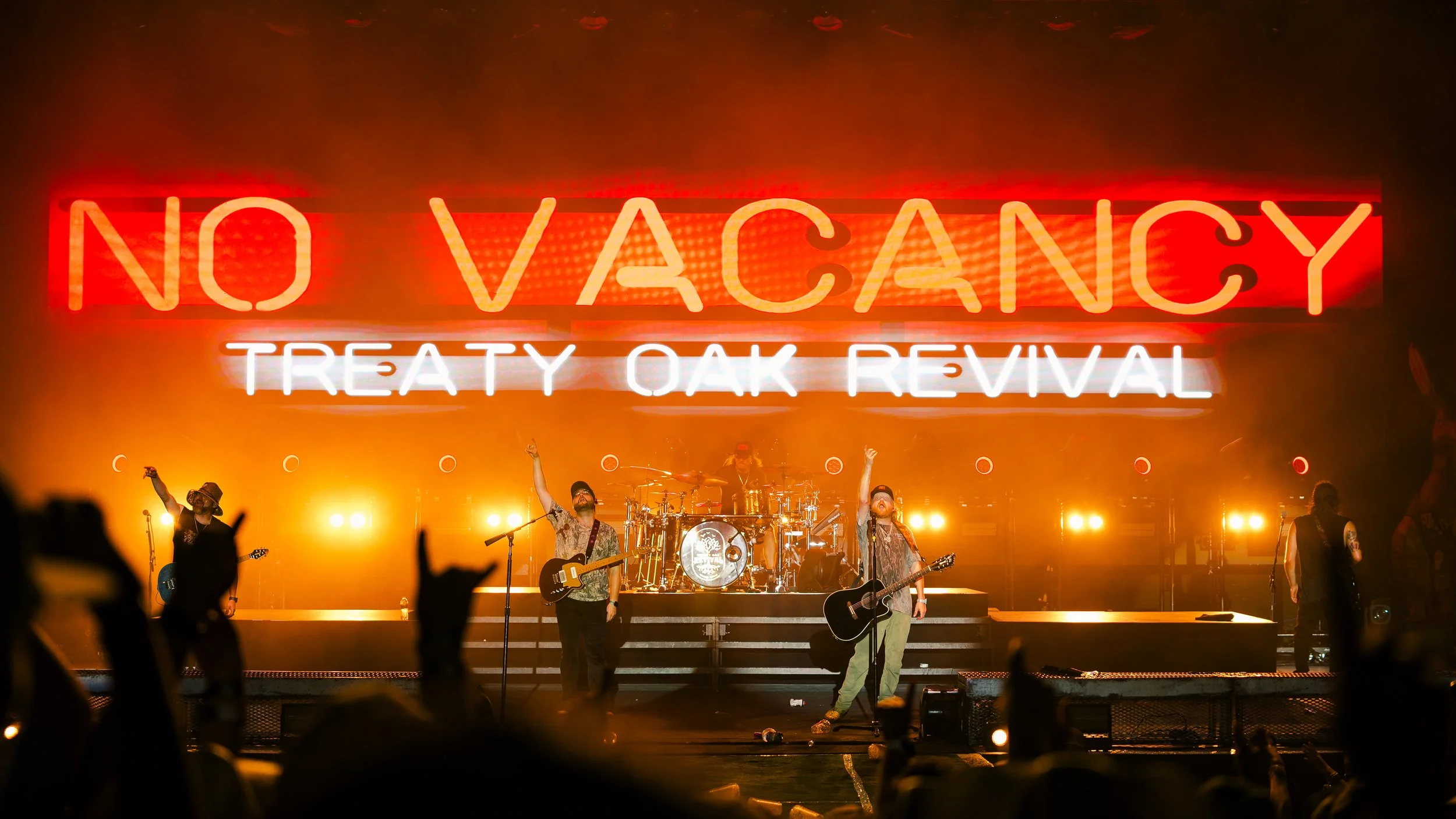 Musicians performing on stage at a concert with a large red and white neon sign reading 'NO VACANCY' and 'TREATY OAK REVIVAL' in the background. Audience silhouettes are visible in the foreground.