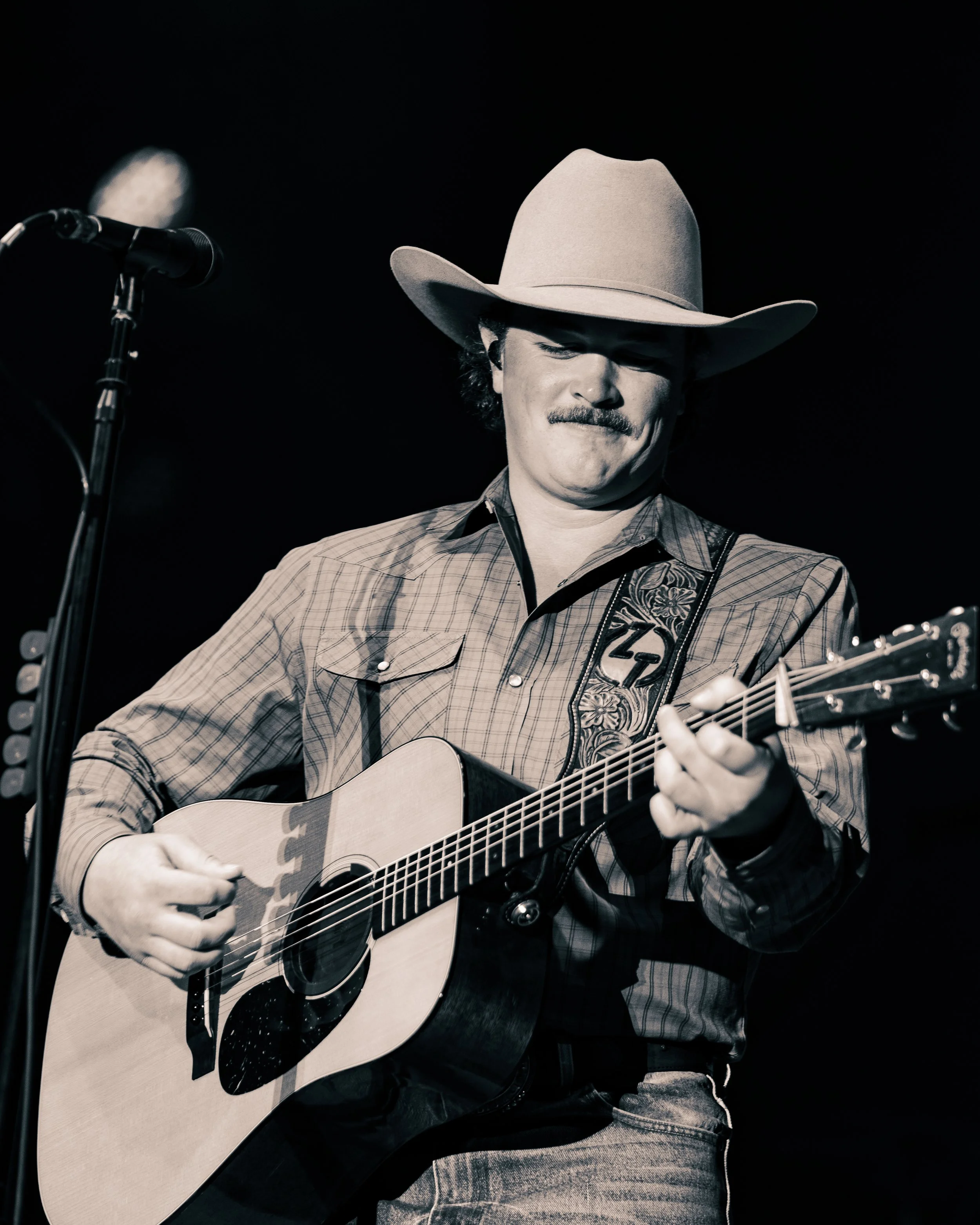 A man wearing a cowboy hat and plaid shirt playing an acoustic guitar on stage with a microphone nearby.