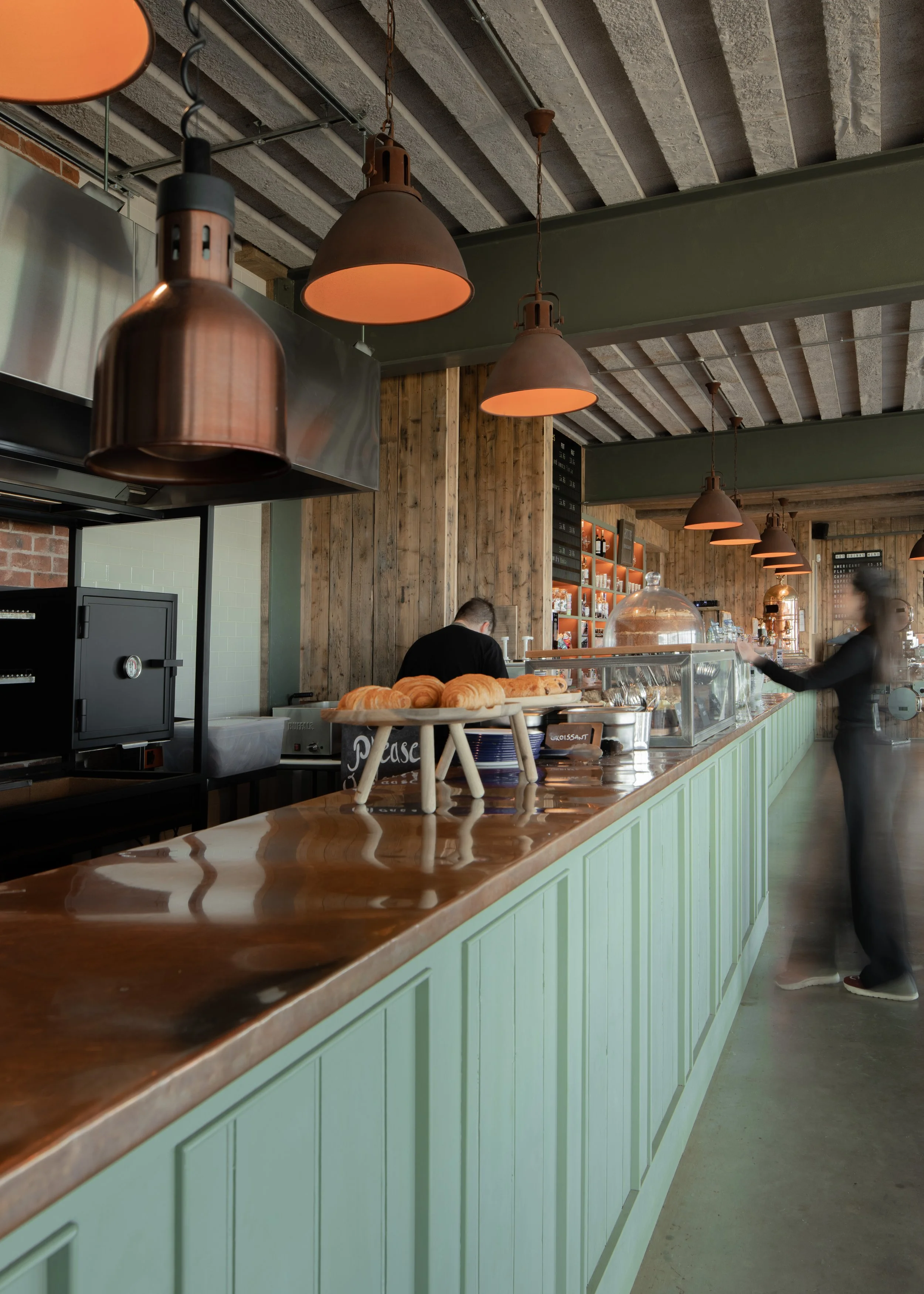 Interior of a modern coffee shop with a wooden counter, hanging copper pendant lights, and wall shelves with various items. A person is serving behind the counter, and croissants are displayed on a small stand.
