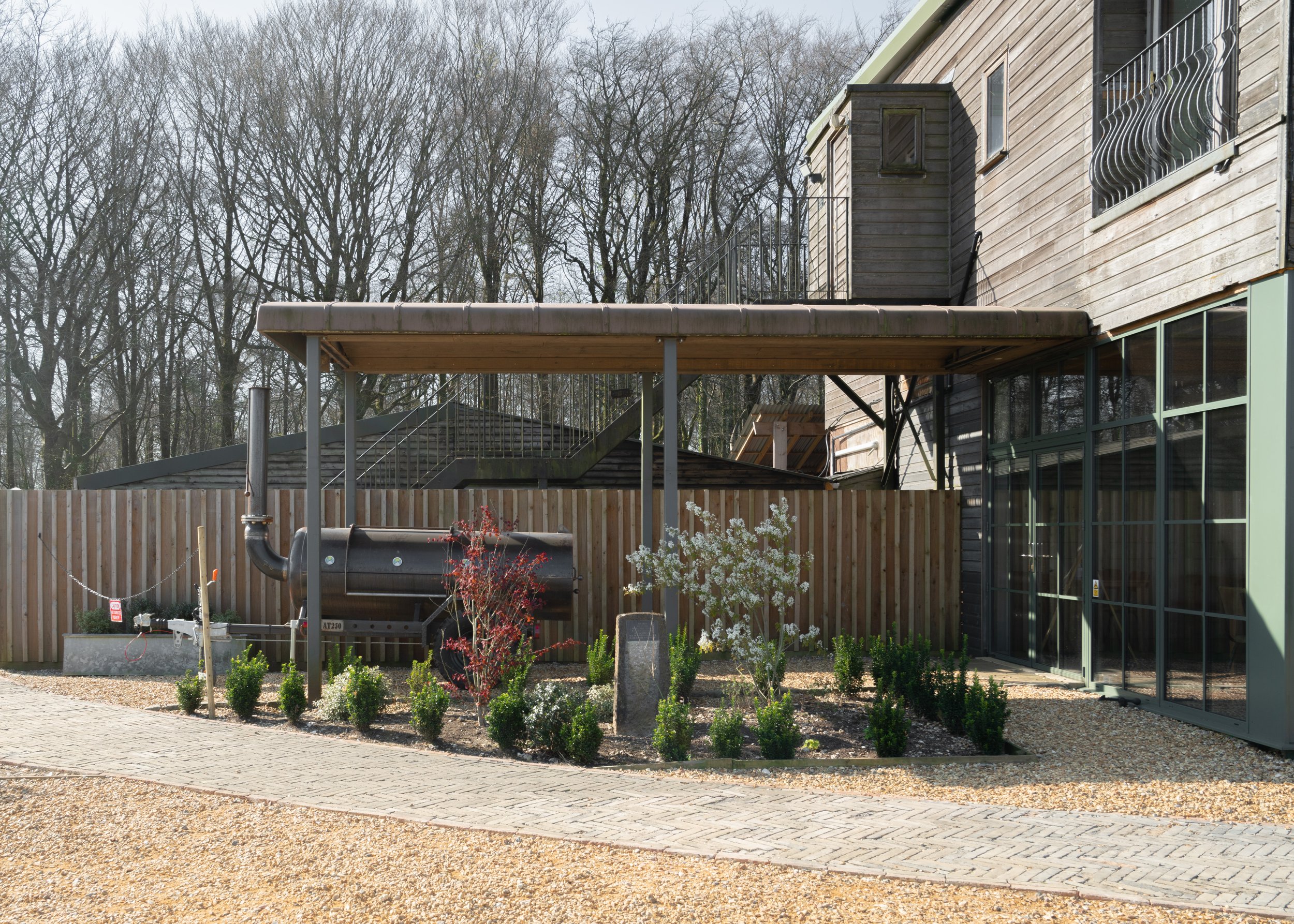 A modern wooden house with large glass windows, an external staircase, a patio with a covered area, and a small garden with bushes and plants, surrounded by leafless trees in the background.