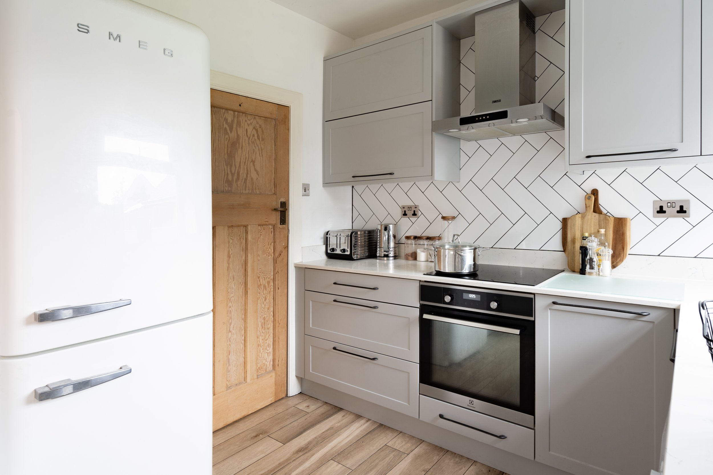 Modern kitchen located in Dorset and photographed by interior photographer Captured Spaces Studio with grey cabinets, white countertop, black stove, stainless steel appliances, wooden cutting board, and herringbone tile backsplash.