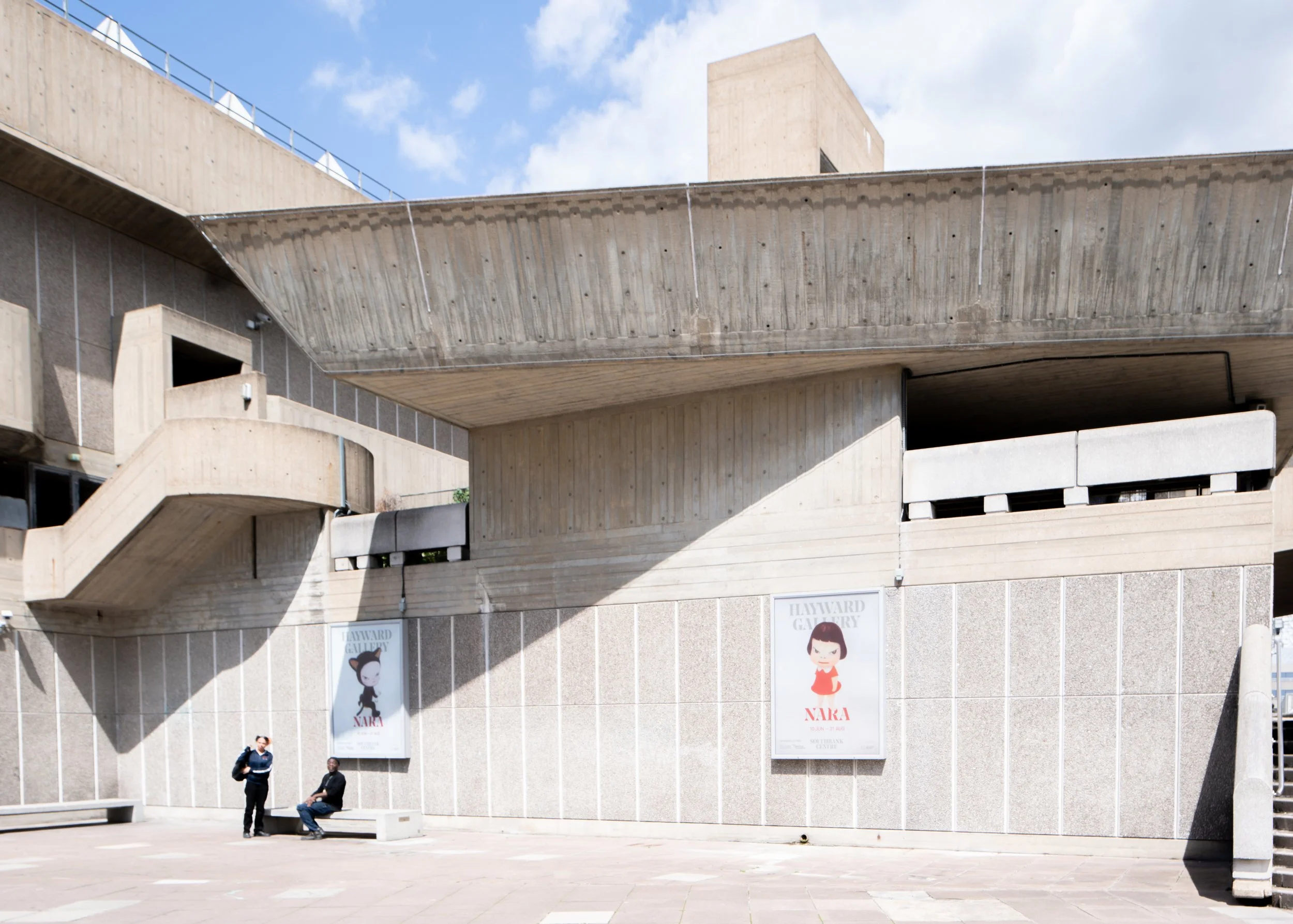 Exterior of the Hayward Gallery with large posters on the wall and a staircase on the left, two people sitting and standing in front of the gallery, blue sky with some clouds.