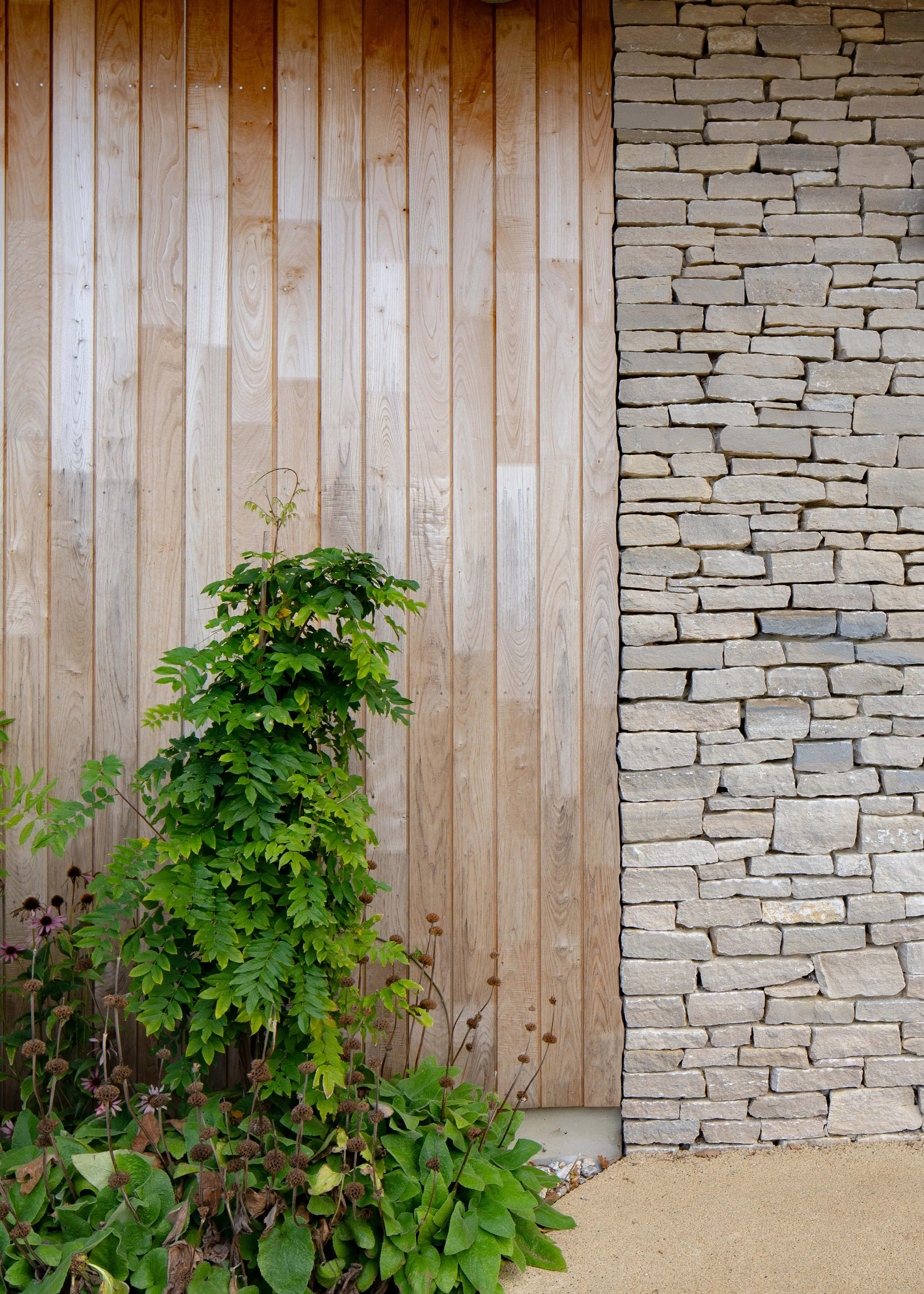 A decorative outdoor wall with a wooden panel on the left and a stone wall on the right, with a green plant and dried flowers at the base.