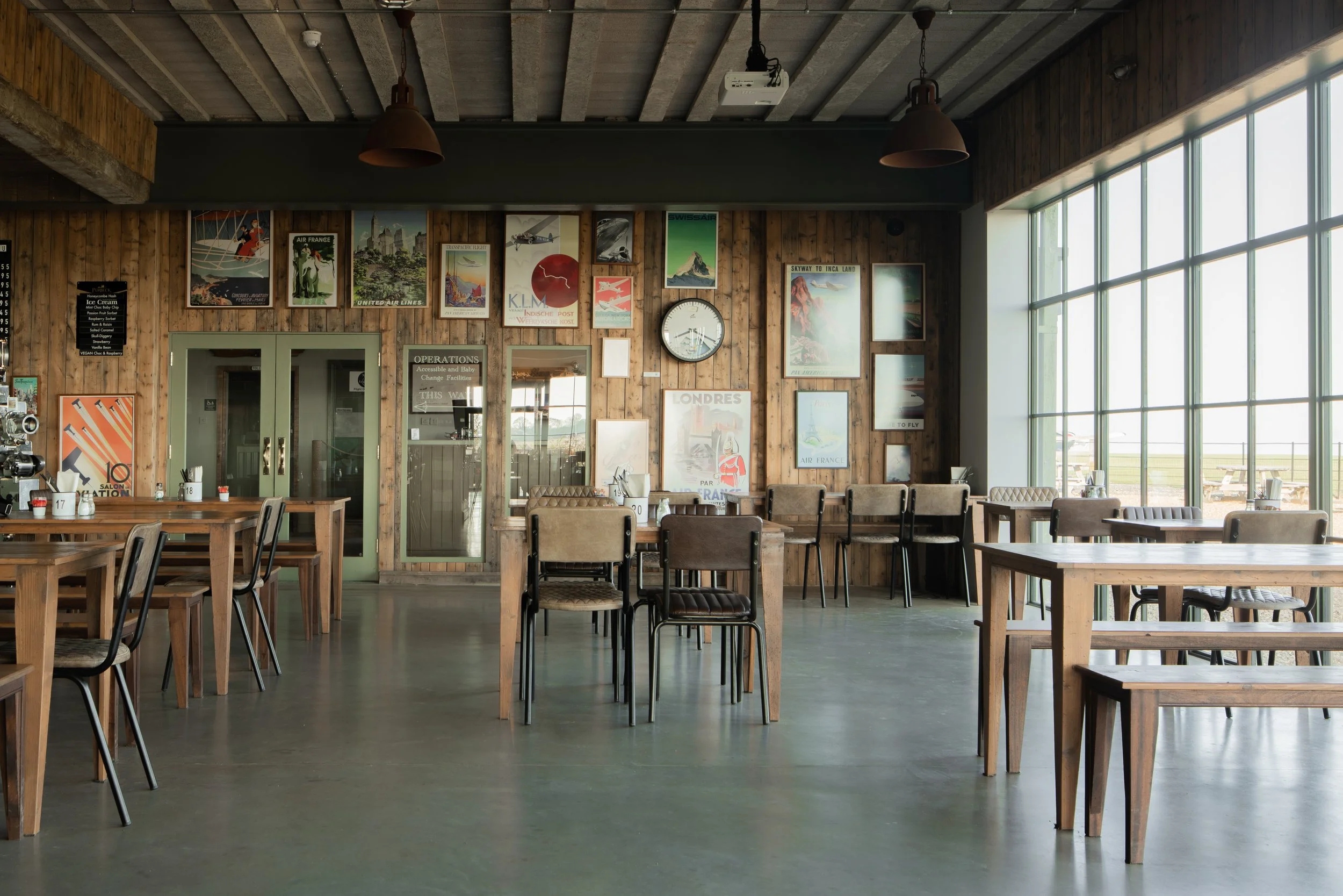Interior of a rustic restaurant or cafe with wooden walls decorated with vintage travel posters, multiple wooden tables and chairs, large windows on the right allowing natural light, and minimal decor.