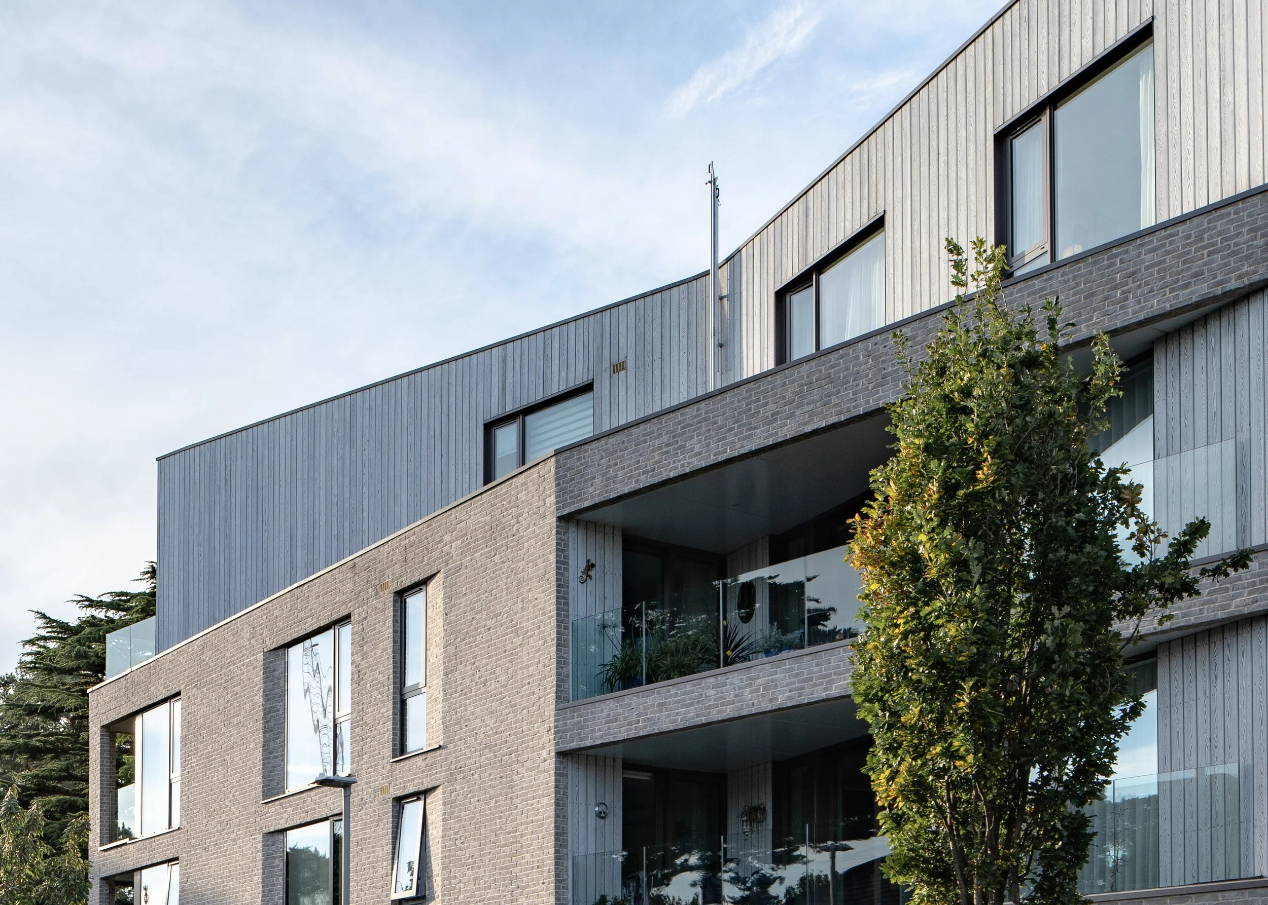 Modern multi-story residential building with large windows and a balcony, partly obscured by a tree, under a partly cloudy sky.