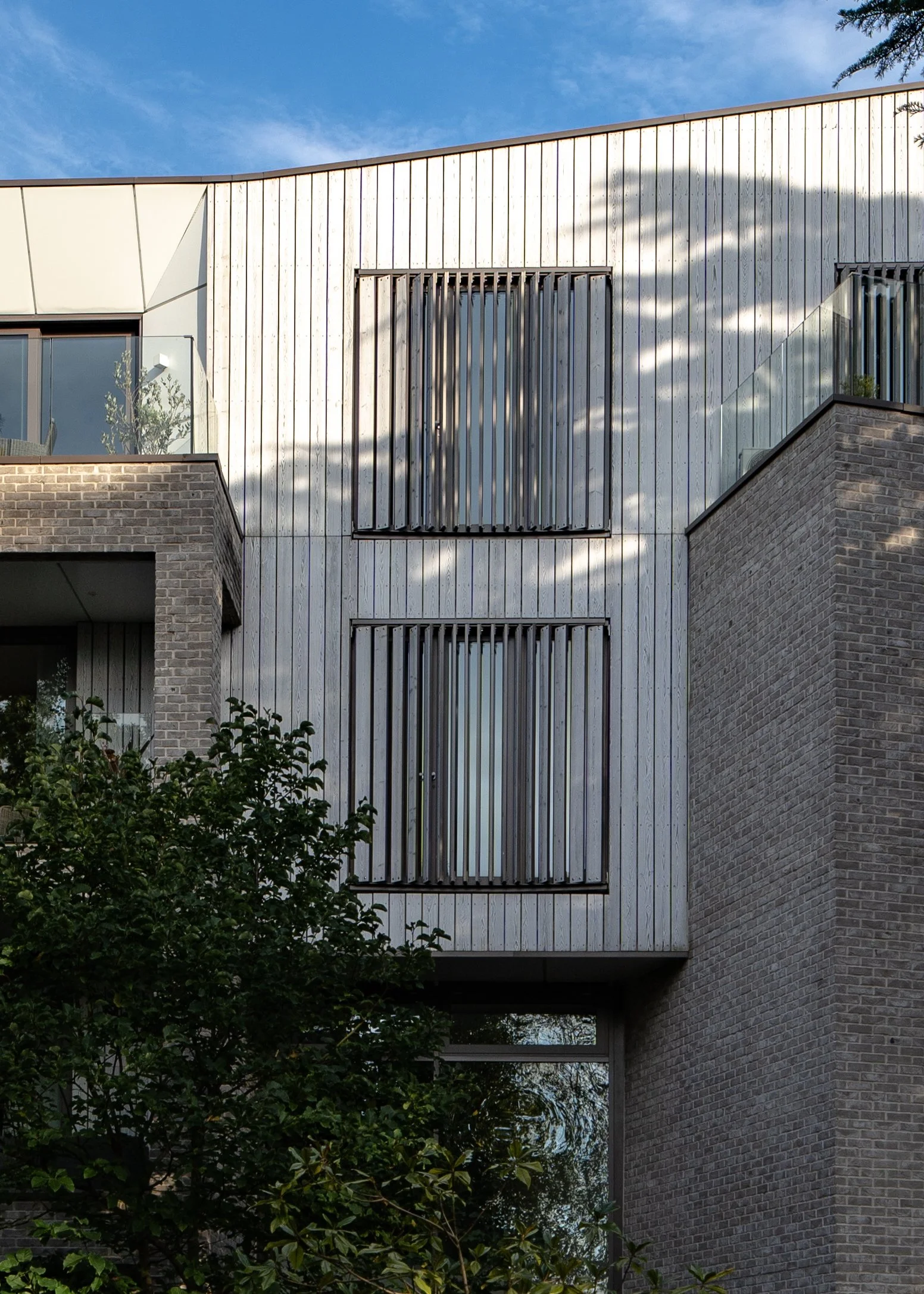 Modern multi-story building located in Dorset and photographed by architectural photographer Captured Spaces Studio with wooden panelling, brick accents, and large windows, with a blue sky and some shadows from trees.
