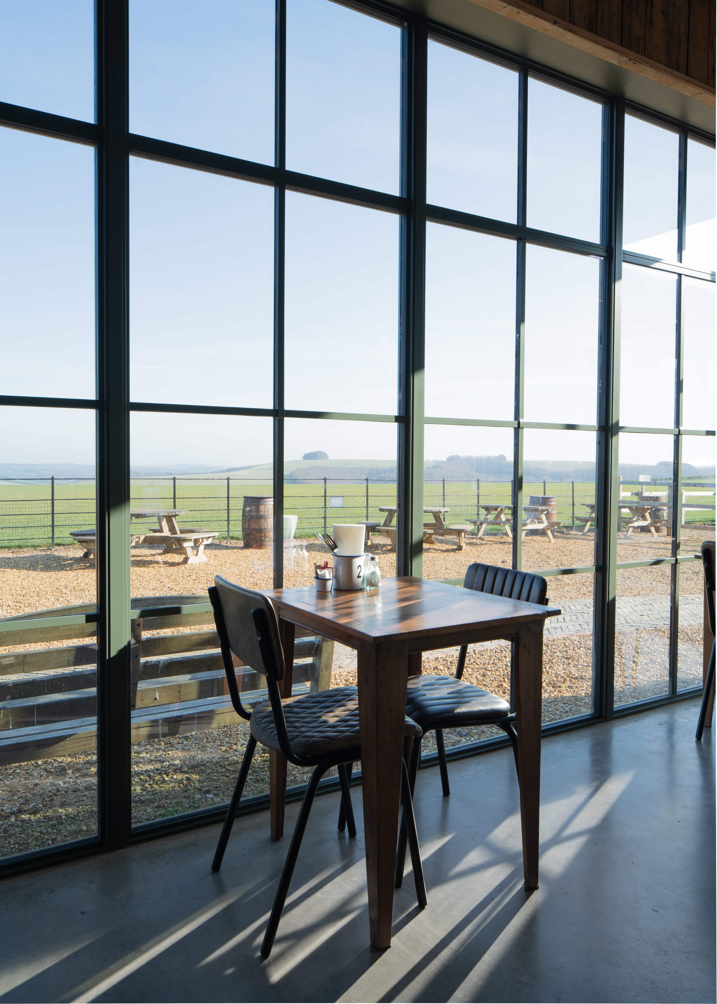 Interior view of a modern restaurant or cafe with large floor-to-ceiling glass windows overlooking a green landscape. There is a wooden table with black chairs, and the sunlight casts shadows on the concrete floor.
