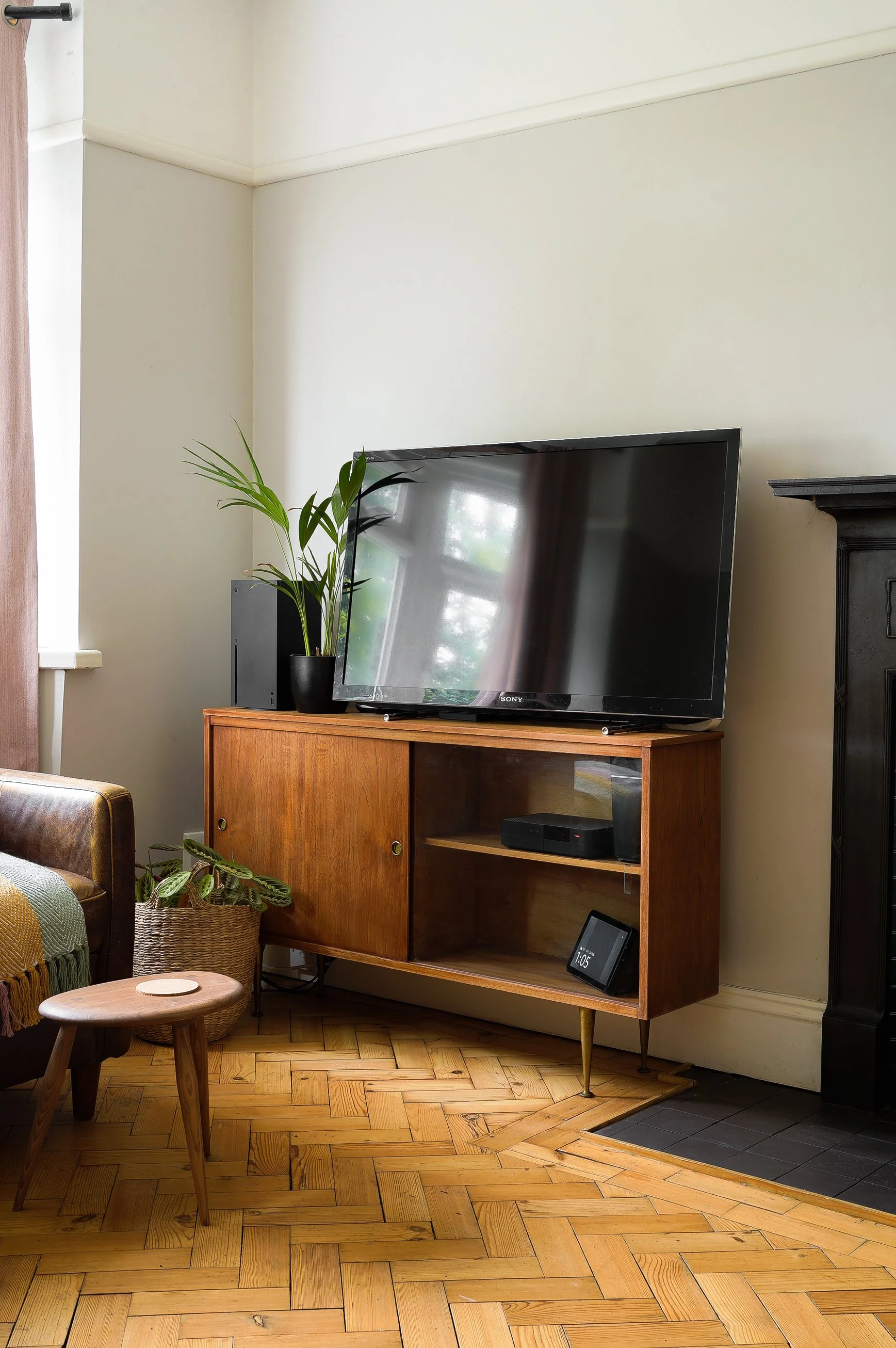 Living room corner with a potted plant, a patterned throw blanket, a small wooden stool, and a fireplace, with hardwood floors and a window with a curtain. Located in Dorset and photographed by interior photographer Captured Spaces Studio