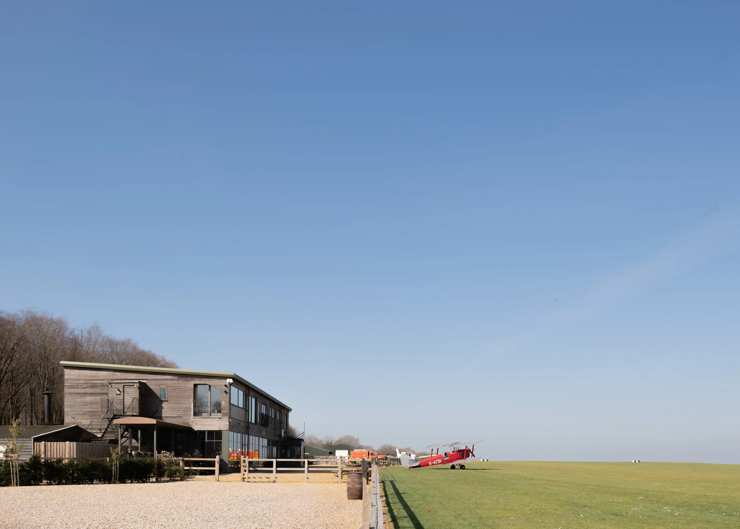 A modern wooden house with large windows sits beside a grassy field. A small airplane with red and white colors is parked on the grass. The sky is clear and blue, with a few trees in the background.