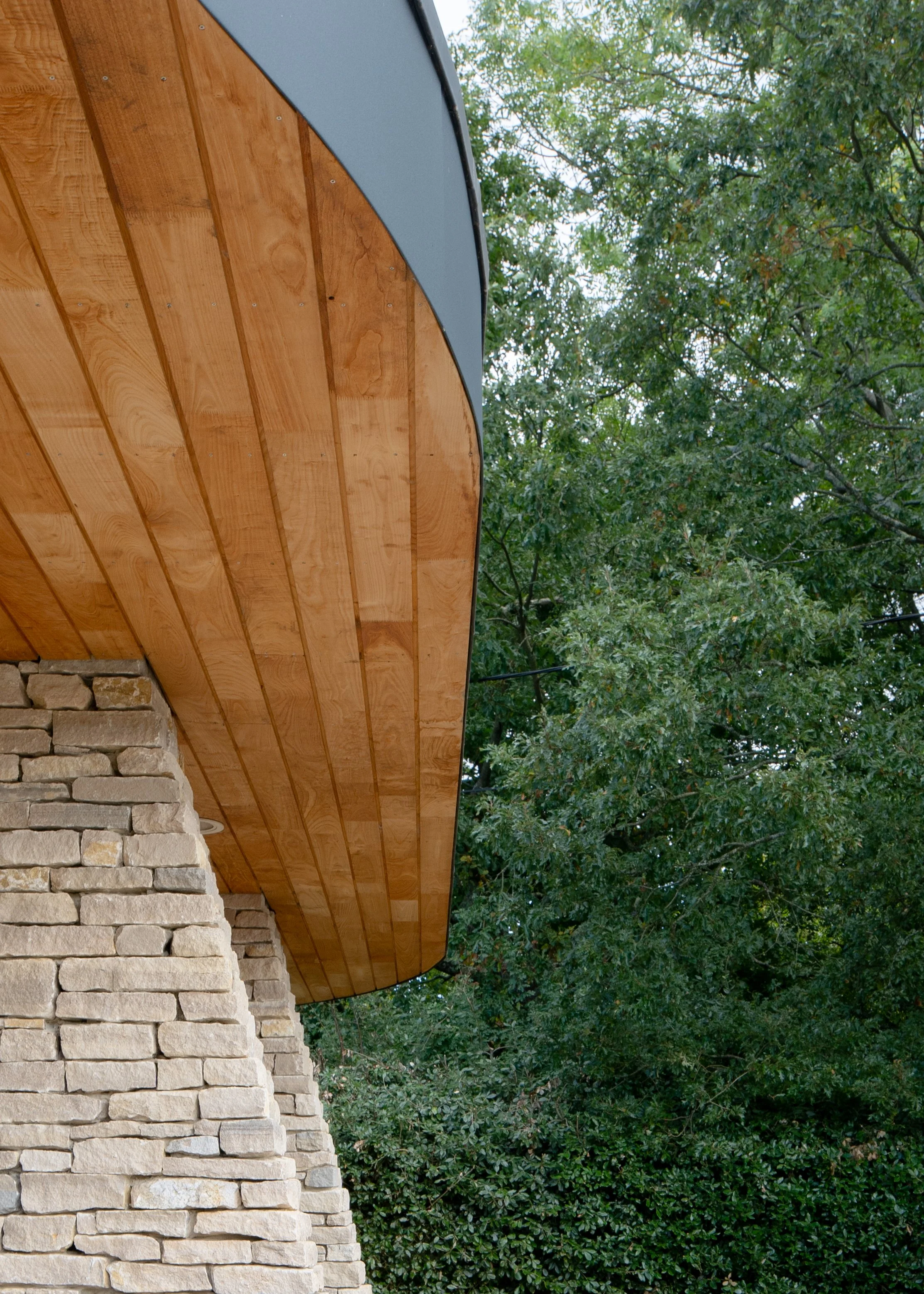 Close-up of a building's exterior, showing a curved wooden roof over a stone brick wall, with green trees in the background.