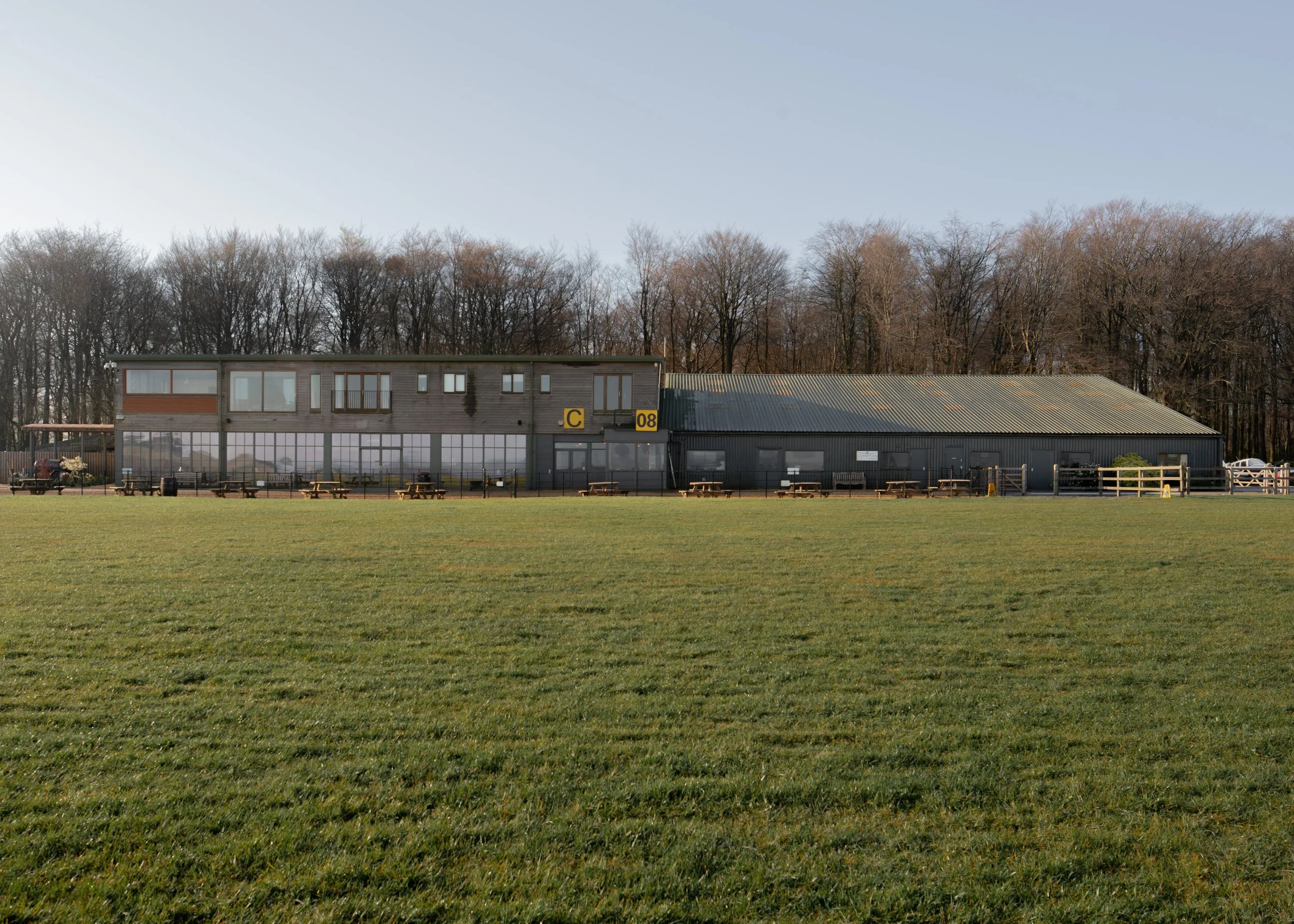 A grassy field with a building in the background, consisting of a mix of glass and metal structures with wooden benches and a line of leafless trees behind it.