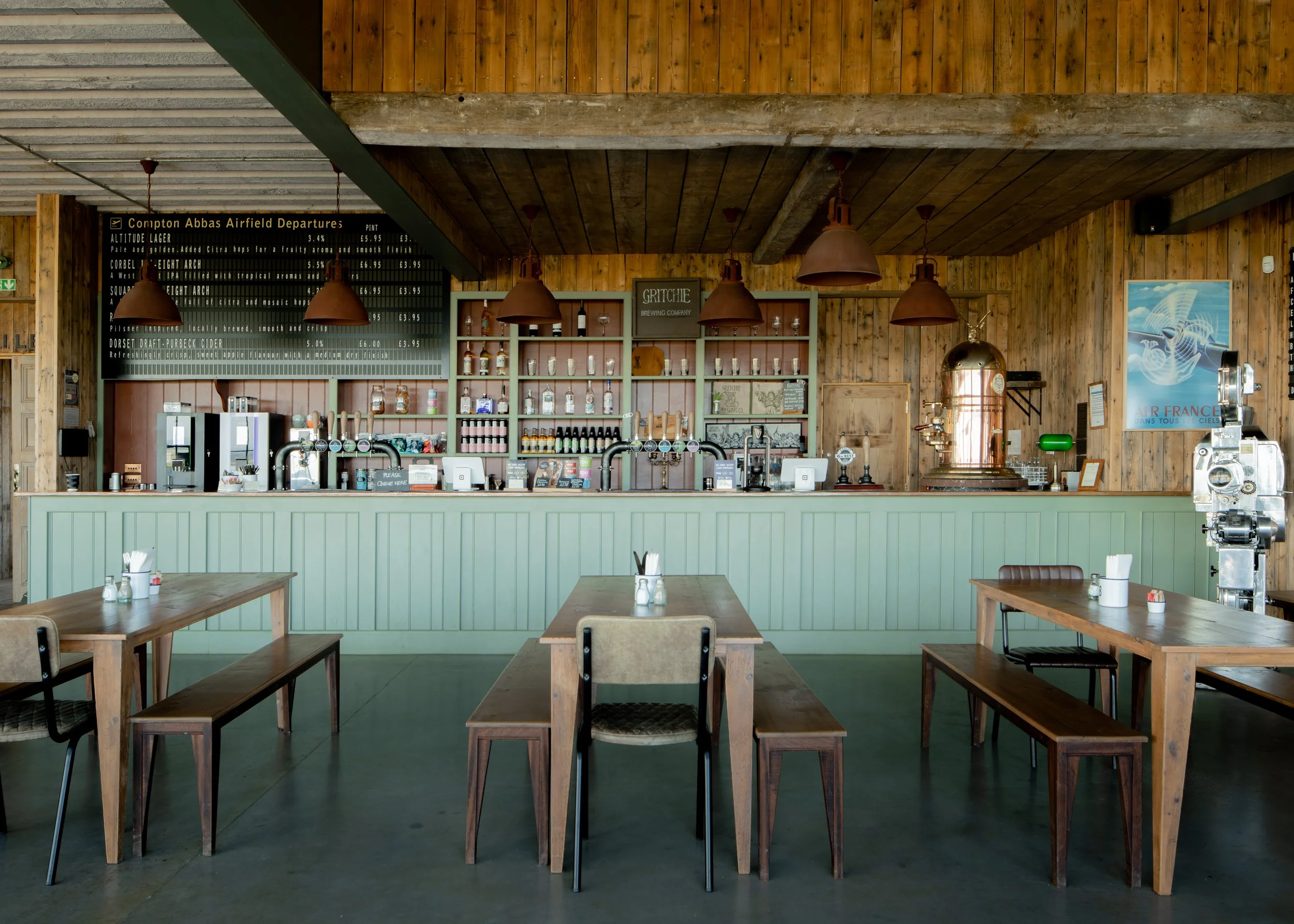 Interior of a rustic cafe with wooden walls and ceiling, featuring a bar area with beer taps, glasses, and a chalkboard menu, and several wooden tables and chairs.