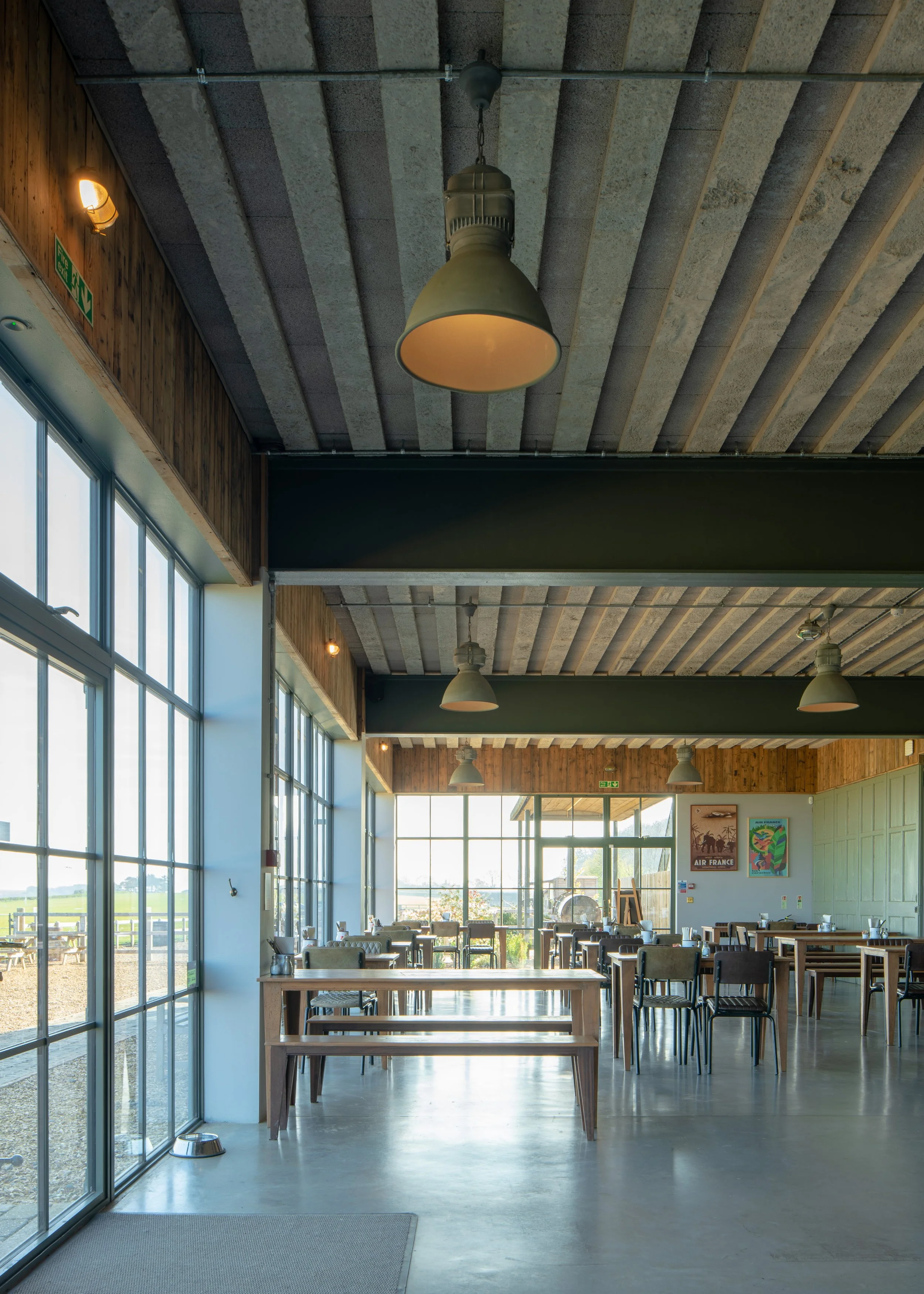 Indoor area with large glass windows, wooden ceiling, hanging industrial light fixtures, and tables and chairs, with daylight streaming in.