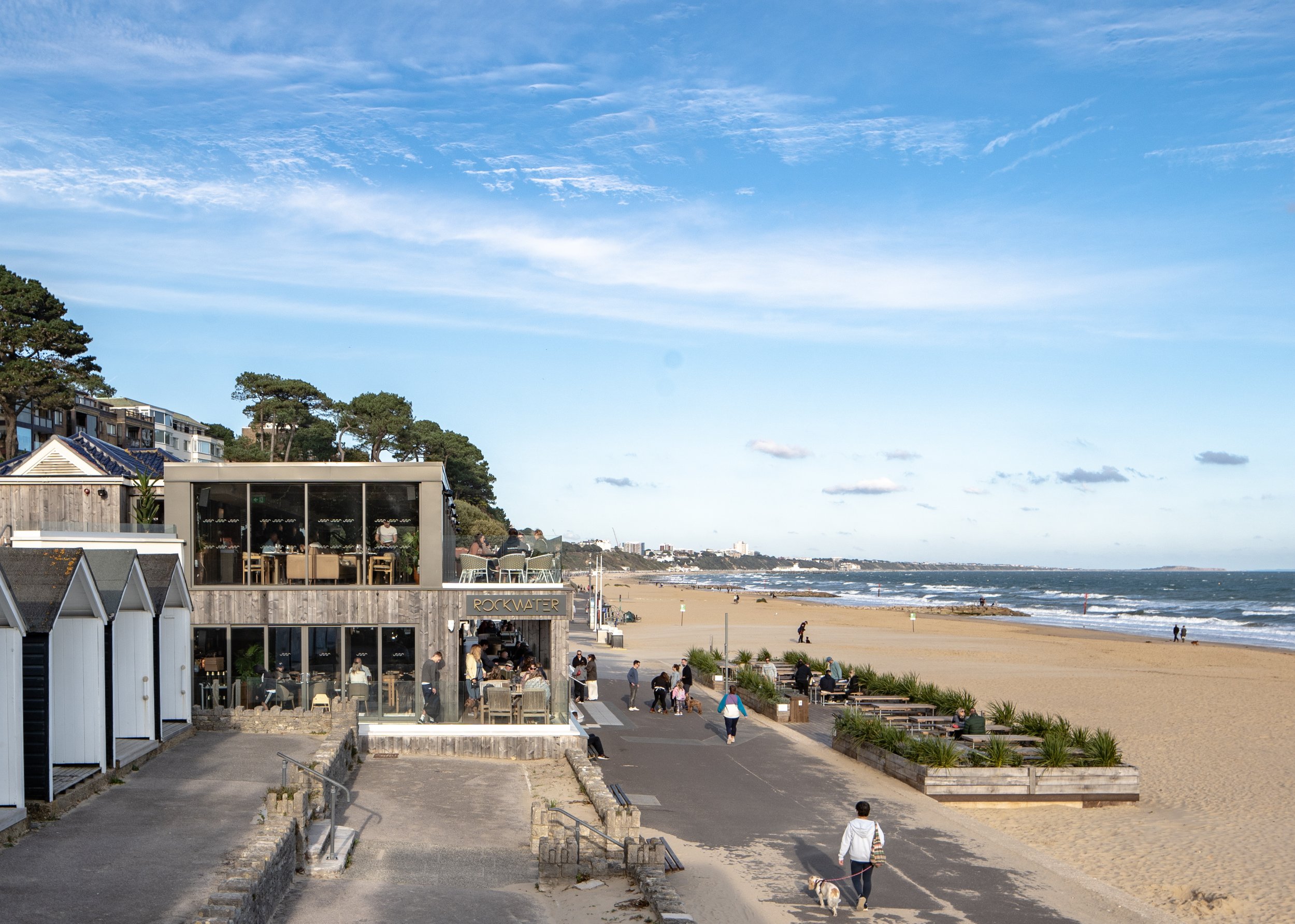 A beachside restaurant named Rockwater with people dining outside on a sunny day, overlooking a sandy beach with some people walking and the ocean in the background.