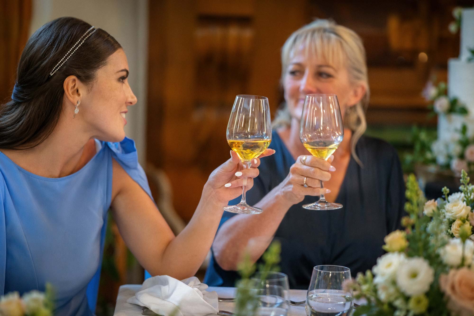 Two women dressed elegantly, holding glasses of white wine, engaging in a conversation at a festive event with floral decorations on the table.