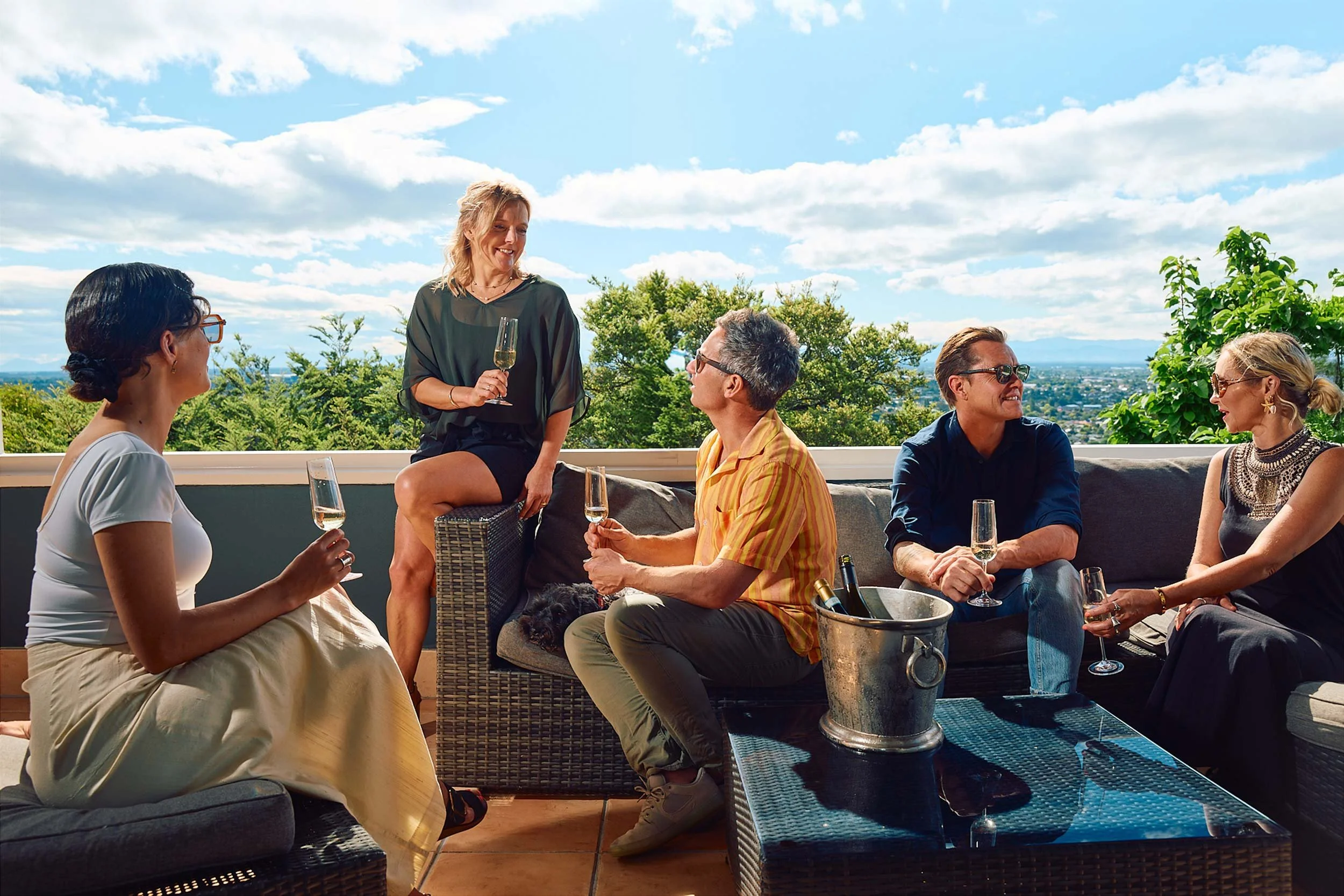 A group of five people socialising outdoors on a sunny patio, holding glasses of champagne with a city view in the background.
