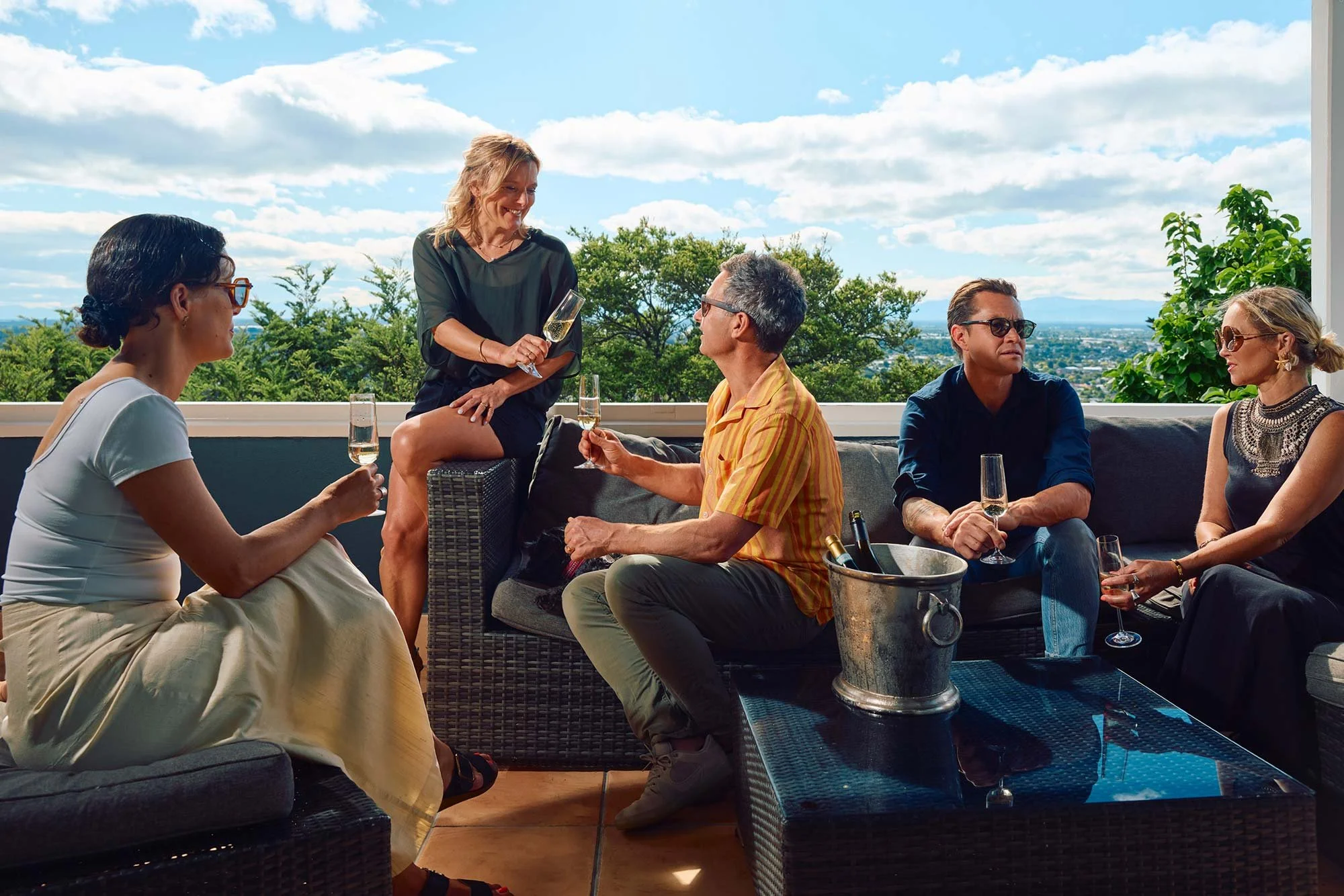 A group of five people socialising, and a couple cheersing with champagne flutes outdoors on a sunny patio, holding glasses of champagne with a city view in the background.