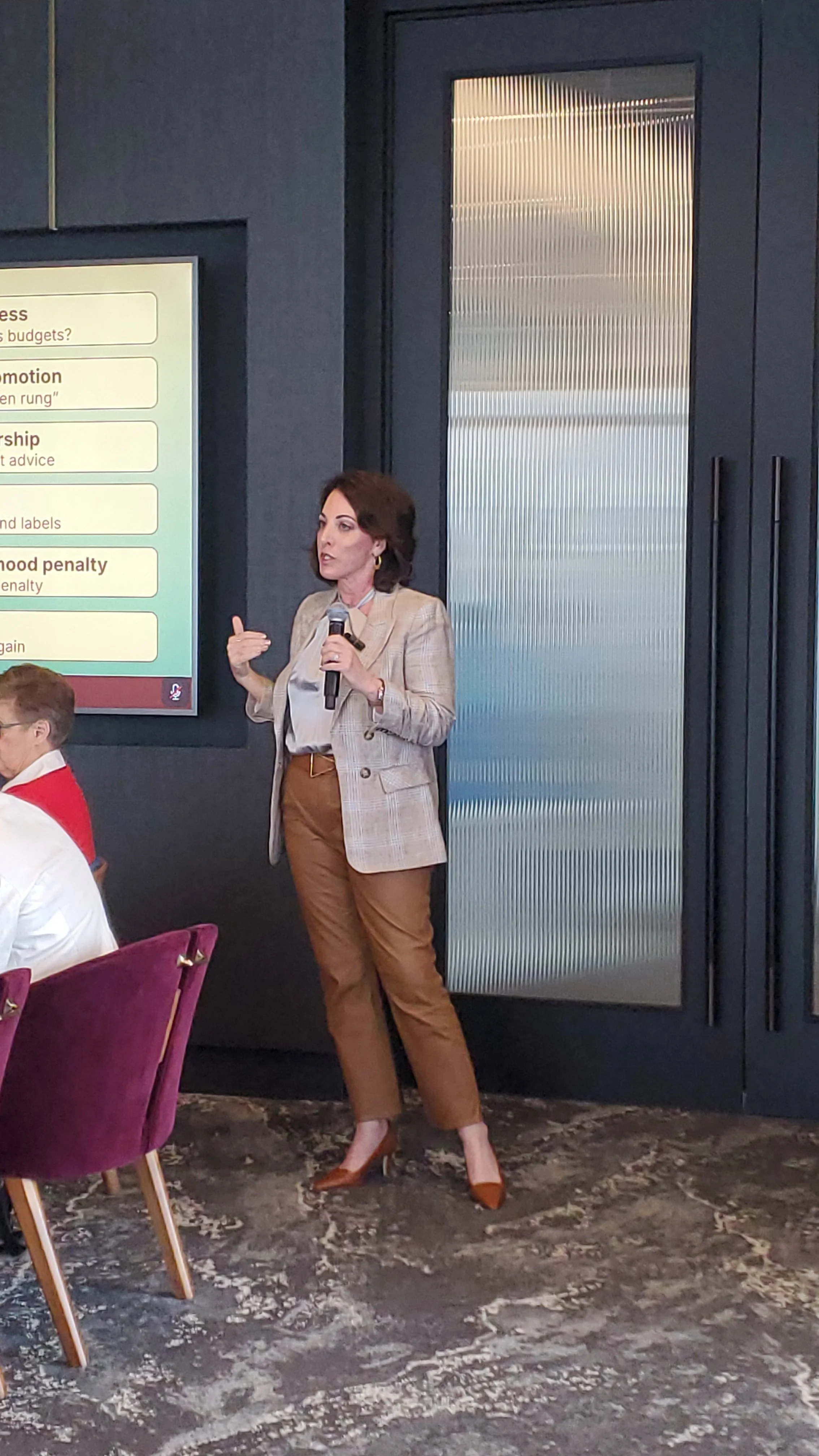 A woman with dark hair wearing a plaid blazer, white blouse, brown pants, and brown heels holding a microphone and speaking at a conference or meeting. A screen with text is visible in the background, and a partially visible person is seated in front