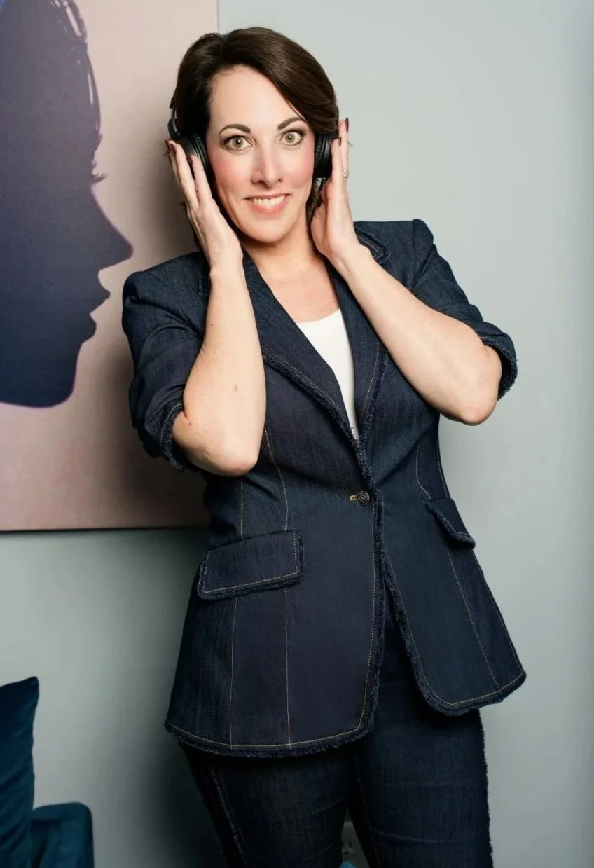 A woman with dark hair wearing a denim blazer and matching pants, holding headphones to her ears and smiling at the camera, standing in front of a wall with a silhouette painting.