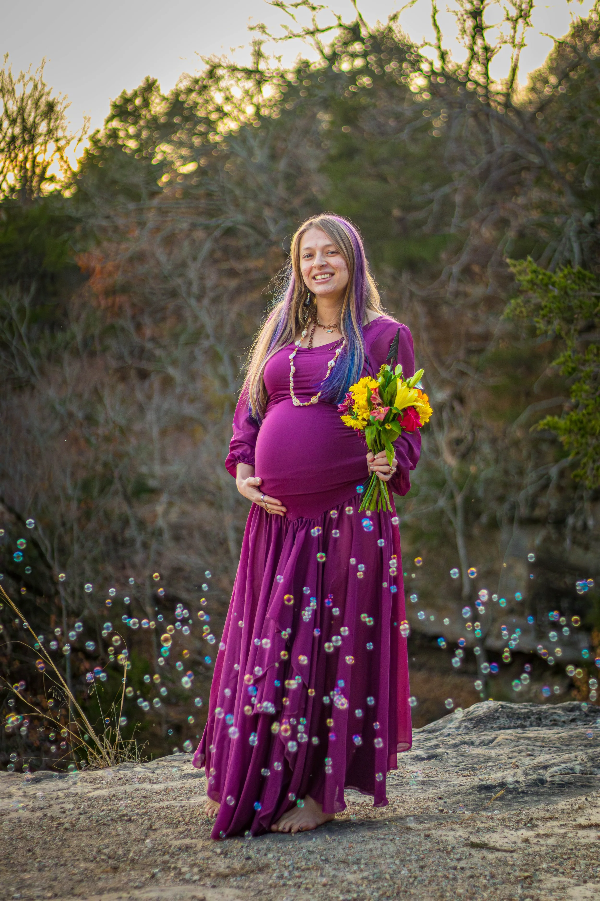 A pregnant woman with colorful hair, dressed in a purple gown, holding a bouquet of yellow and red flowers, standing outdoors on a rock with a nature background.