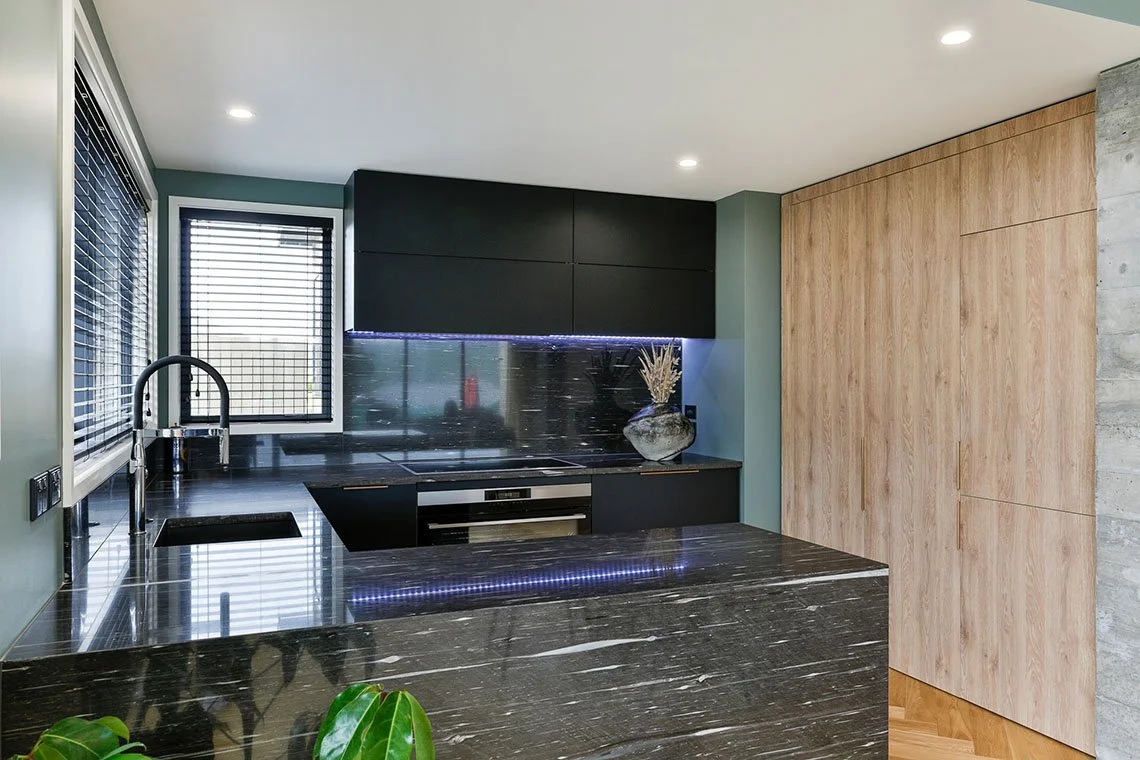 Modern kitchen with black countertops, black cabinetry, a black backsplash, a window with blinds, built-in oven, and wood paneling on the right wall.