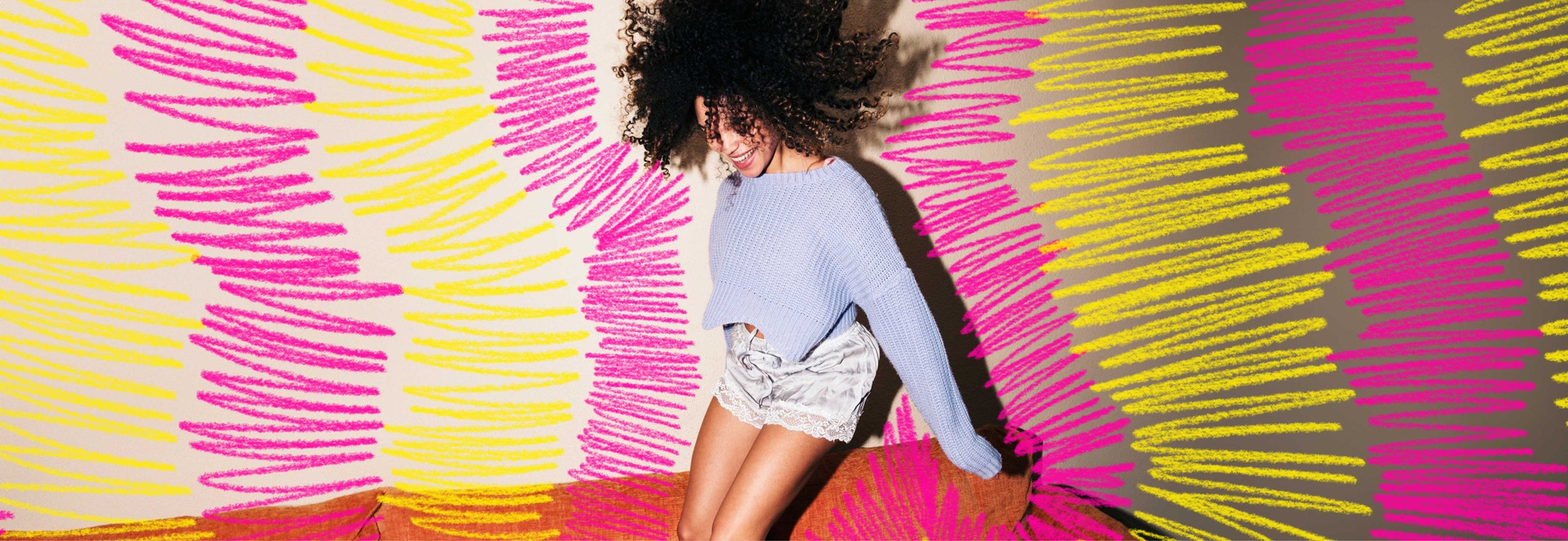 A person with voluminous curly hair and a joyful expression jumps on a couch, wearing a lavender sweater and silky lace-trimmed shorts. Hand-drawn pink and yellow brushstrokes surround them, adding a sense of energy and motion.