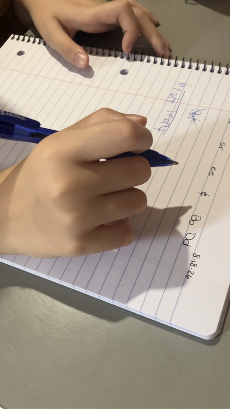 A student writing in a notebook with a blue pen, with handwritten spelling practice on lined paper.