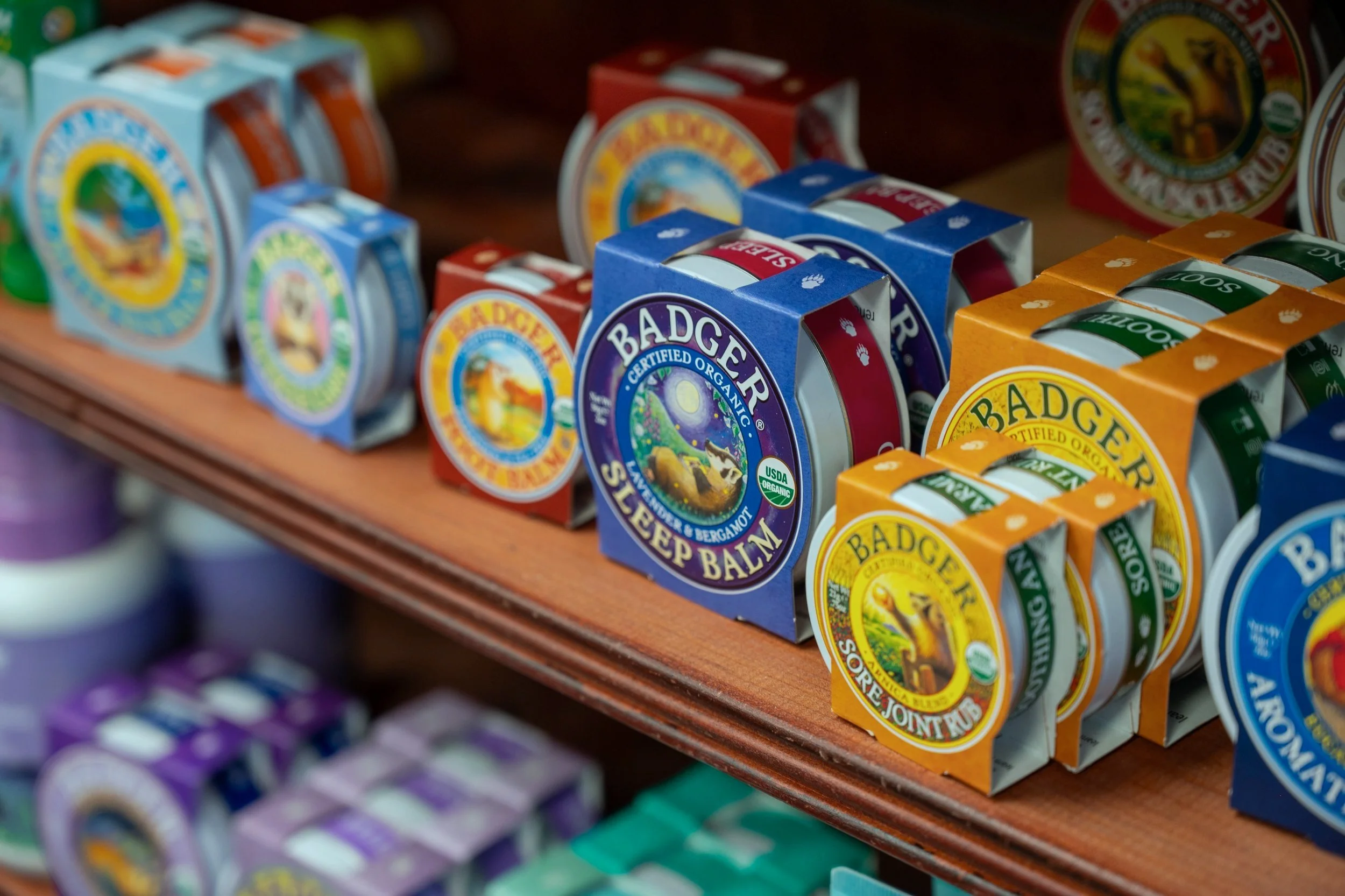 Display of Badger brand balms and salves on a wooden shelf, featuring various colored packaging including blue, orange, red, and purple.