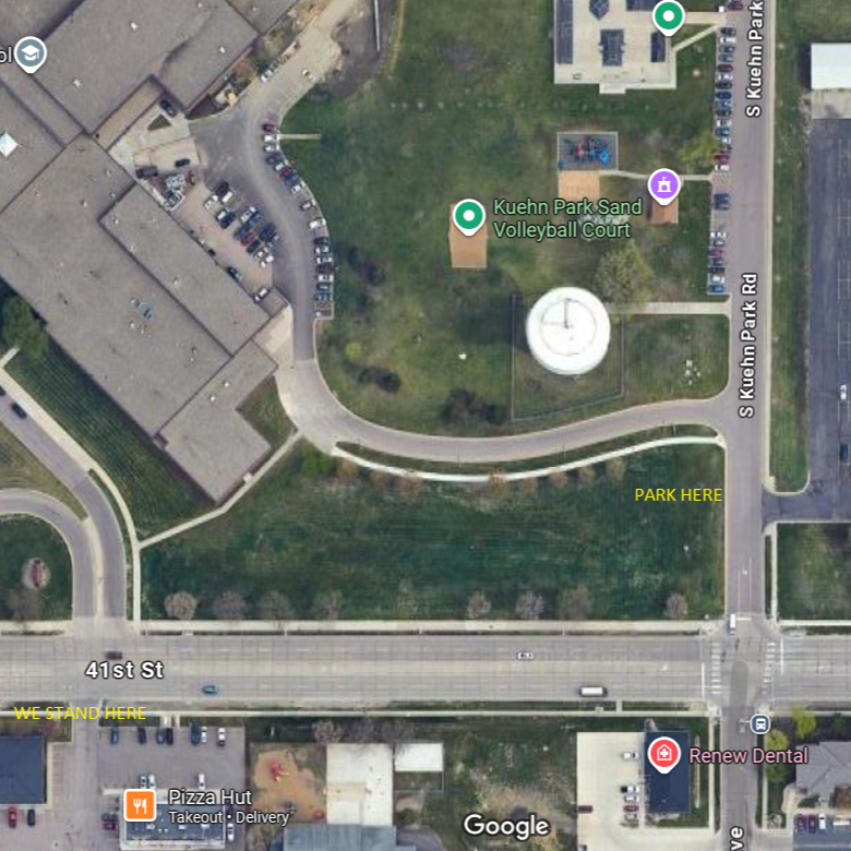 Satellite view of a park named Kuehn Park with a sand volleyball court, a playground, and a picnic area. There is a sidewalk looping around parts of the park, with a marked parking area and a sign indicating "PARK HERE." Adjacent to the park is a street labeled 41st St, and nearby businesses include a pizza restaurant and a dental office.
