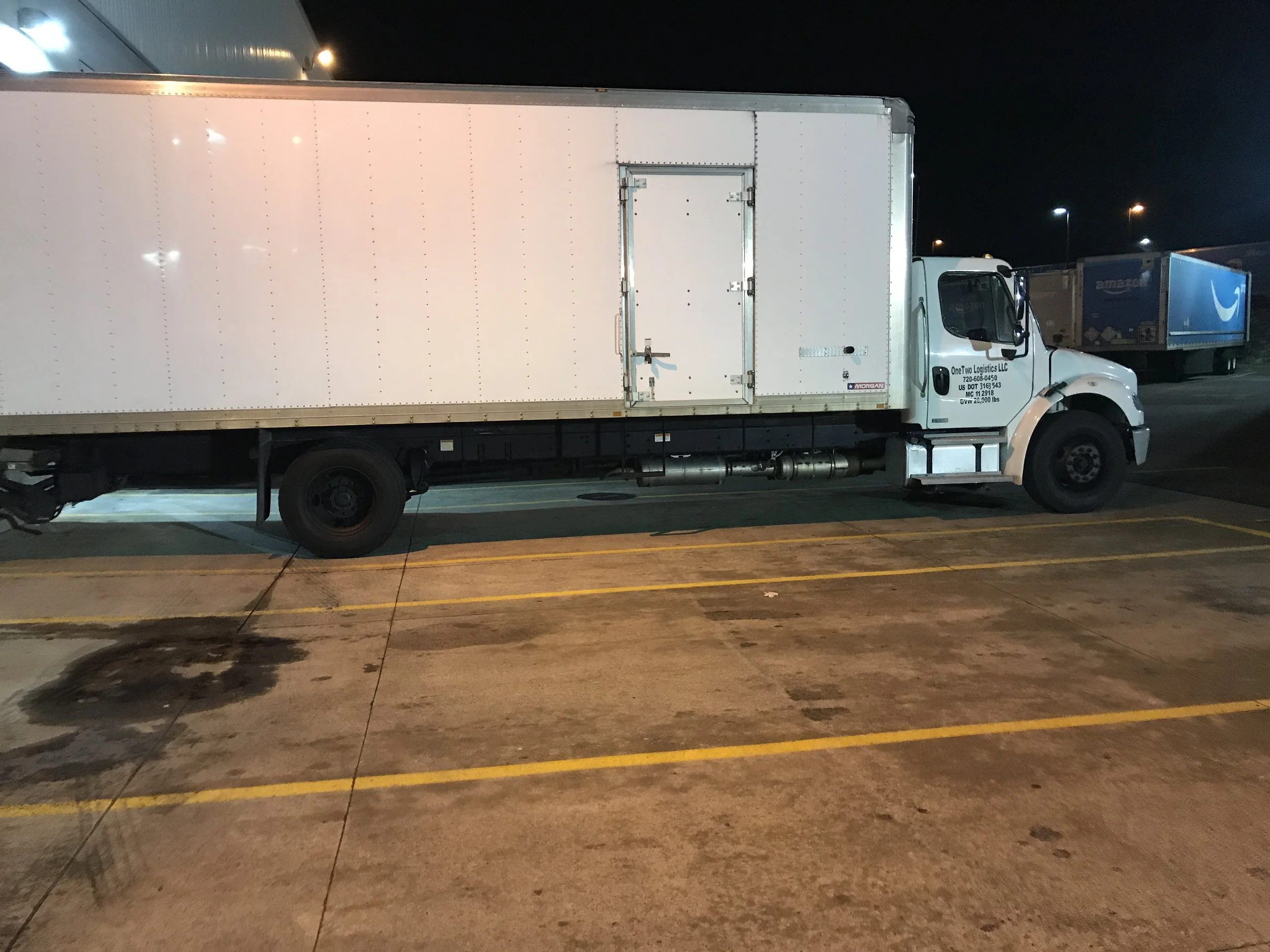 A white box truck parked in a parking lot at night, with another truck visible in the background.