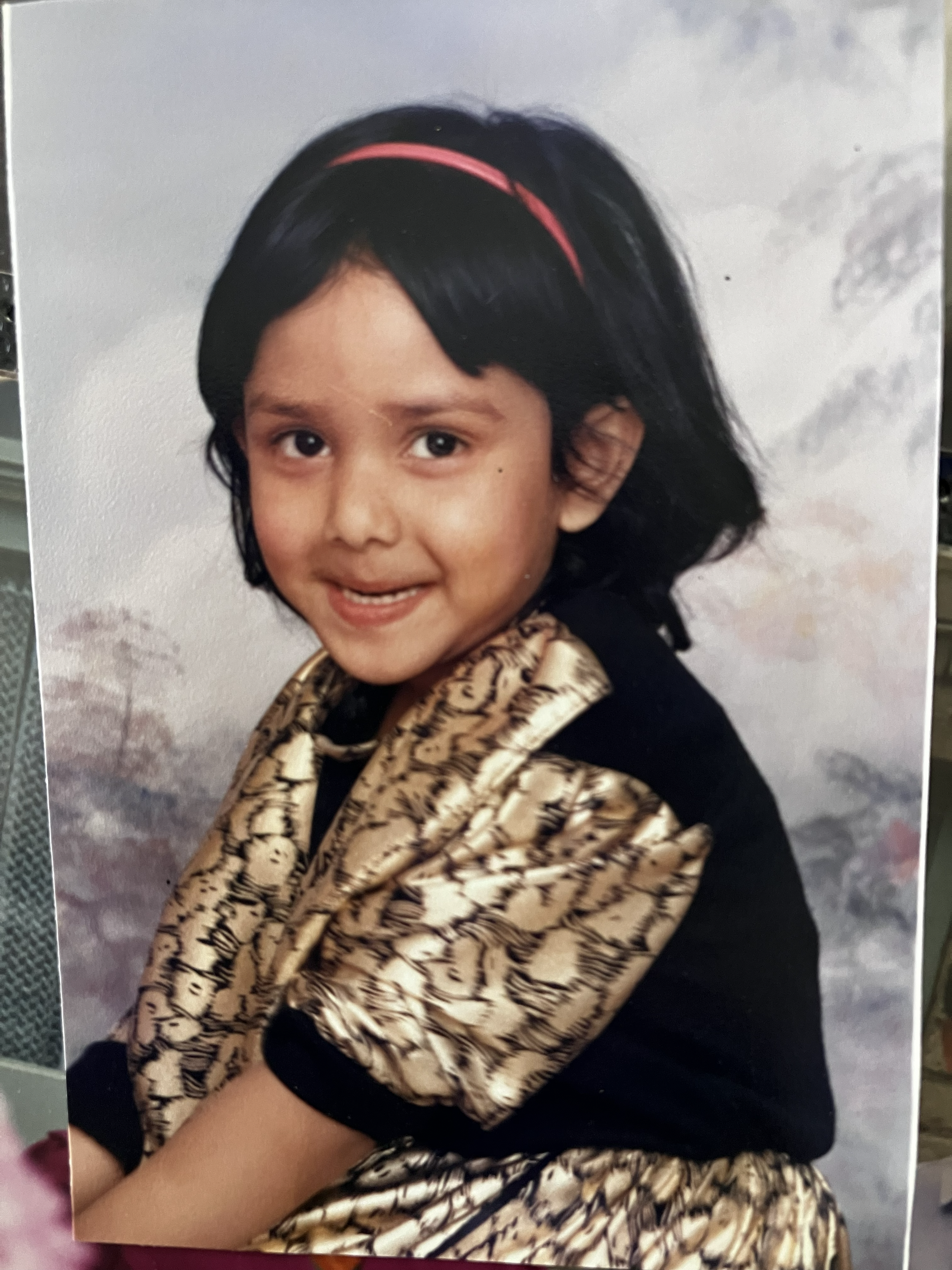 A young girl with short black hair, wearing a pink headband, smiling and looking at the camera. She is dressed in a black shirt with a beige and black patterned scarf or shawl. The background appears to be a cloudy sky.