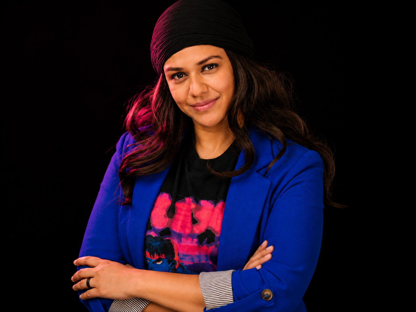 A woman with long wavy hair wearing a black headscarf, a bright blue blazer, and a black T-shirt with a colorful graphic design, standing against a black background.