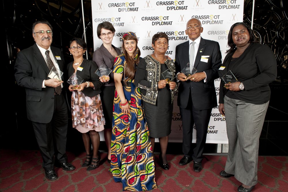 Group of eight diverse individuals posing with glass awards in front of a backdrop that reads 'Grassroot Diplomat'.