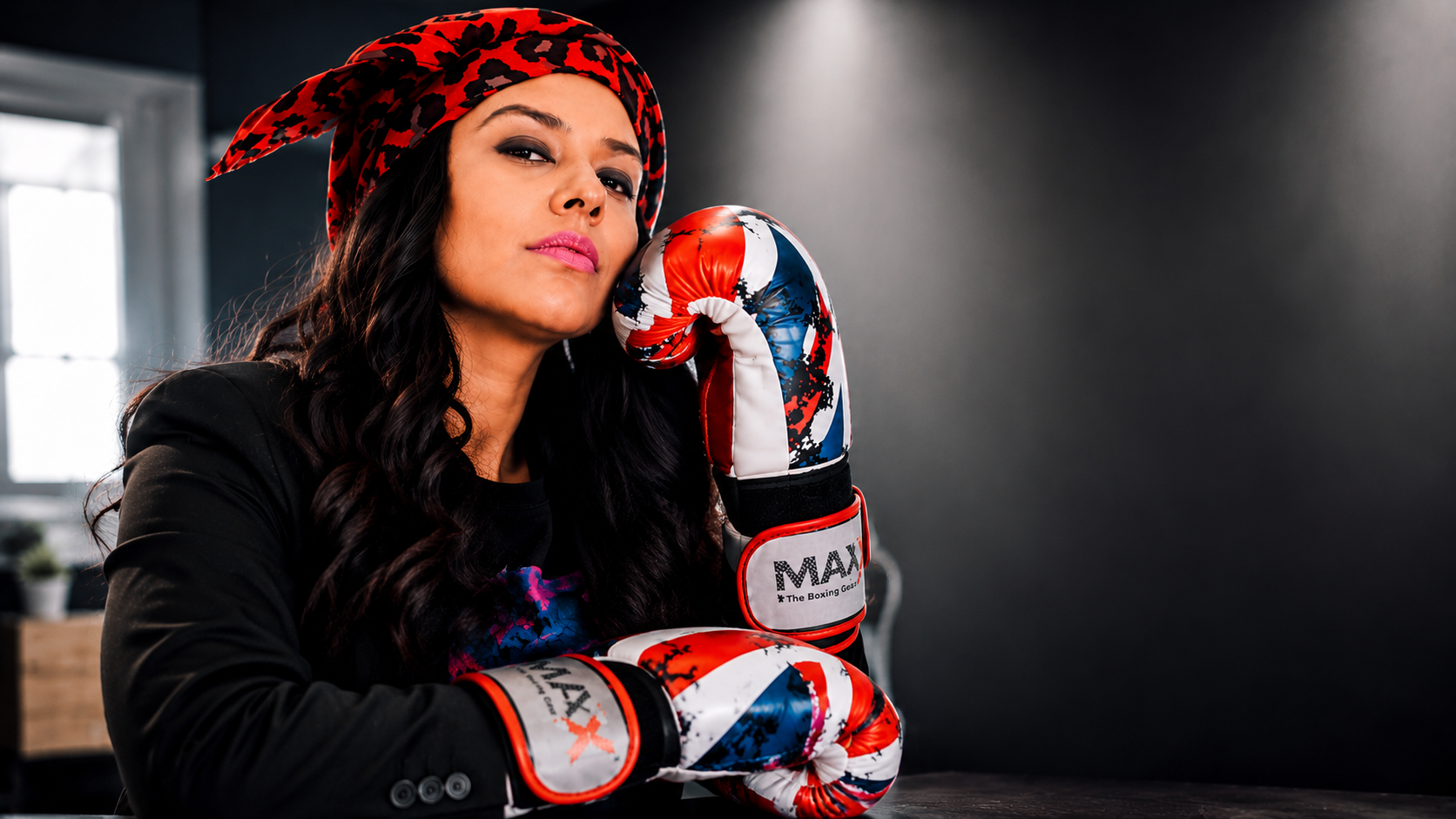 A woman wearing boxing gloves and a red leopard print headscarf, resting her chin on her boxing glove in a gym setting.
