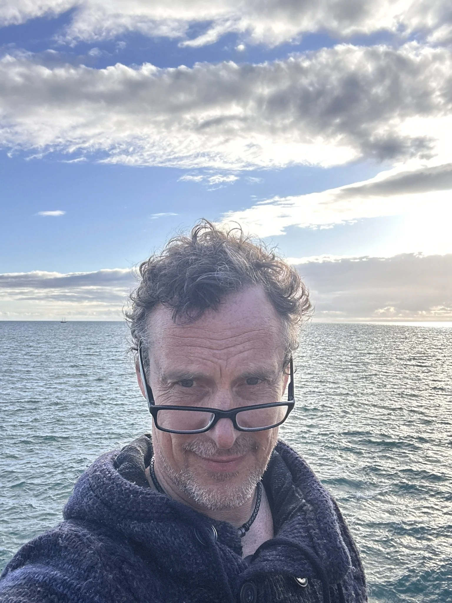 Ein Mann mit Brille macht ein Selfie vor dem Meer bei Sonnenuntergang.