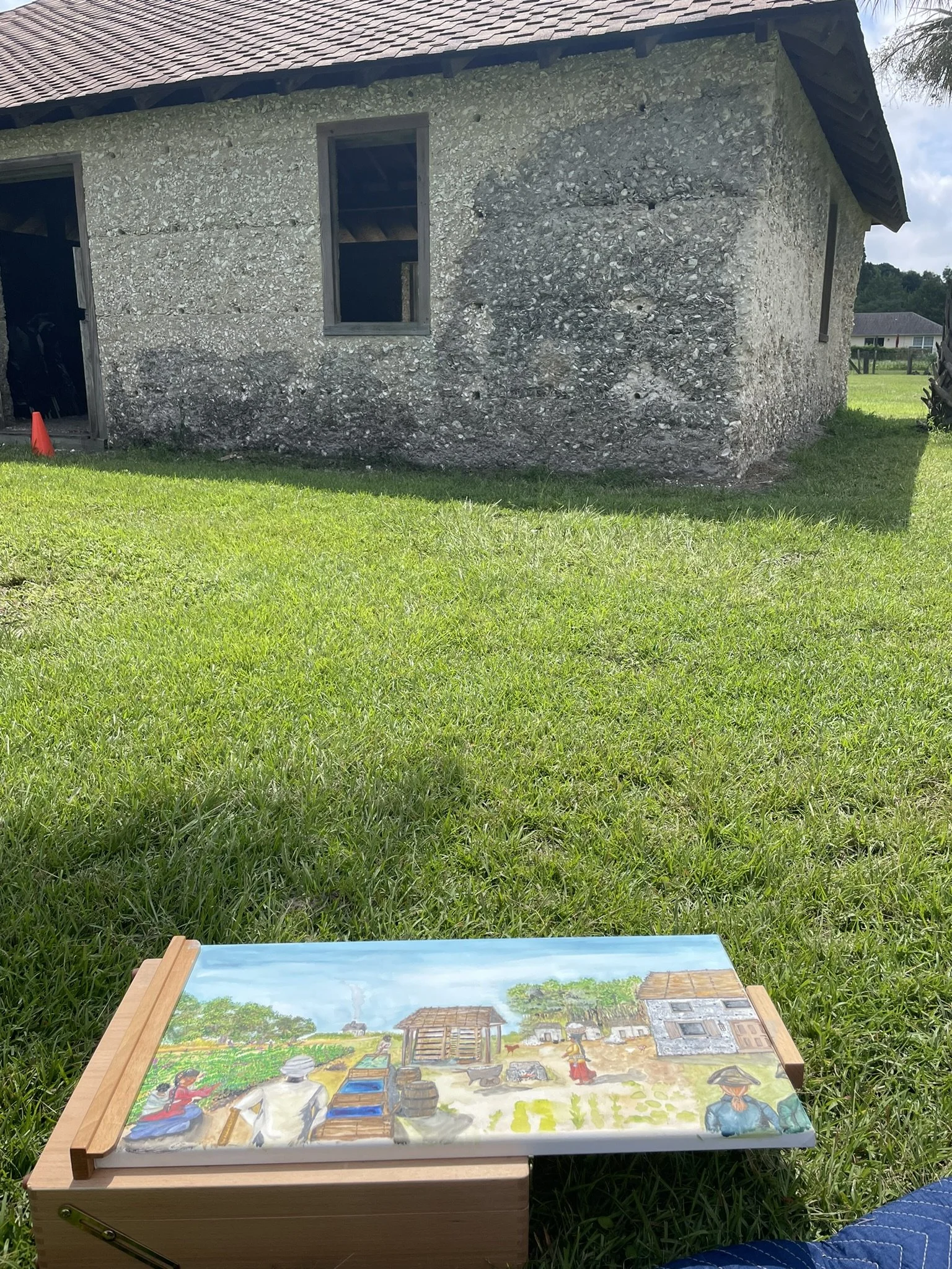 Dr. Katherine Seeber sits on the ground with a canvas in her lap, paint and brushes to the right. In front of her is the Barnwell Family Tabby. You can see she is painting the Tabby in a historical interpretation illustration. 