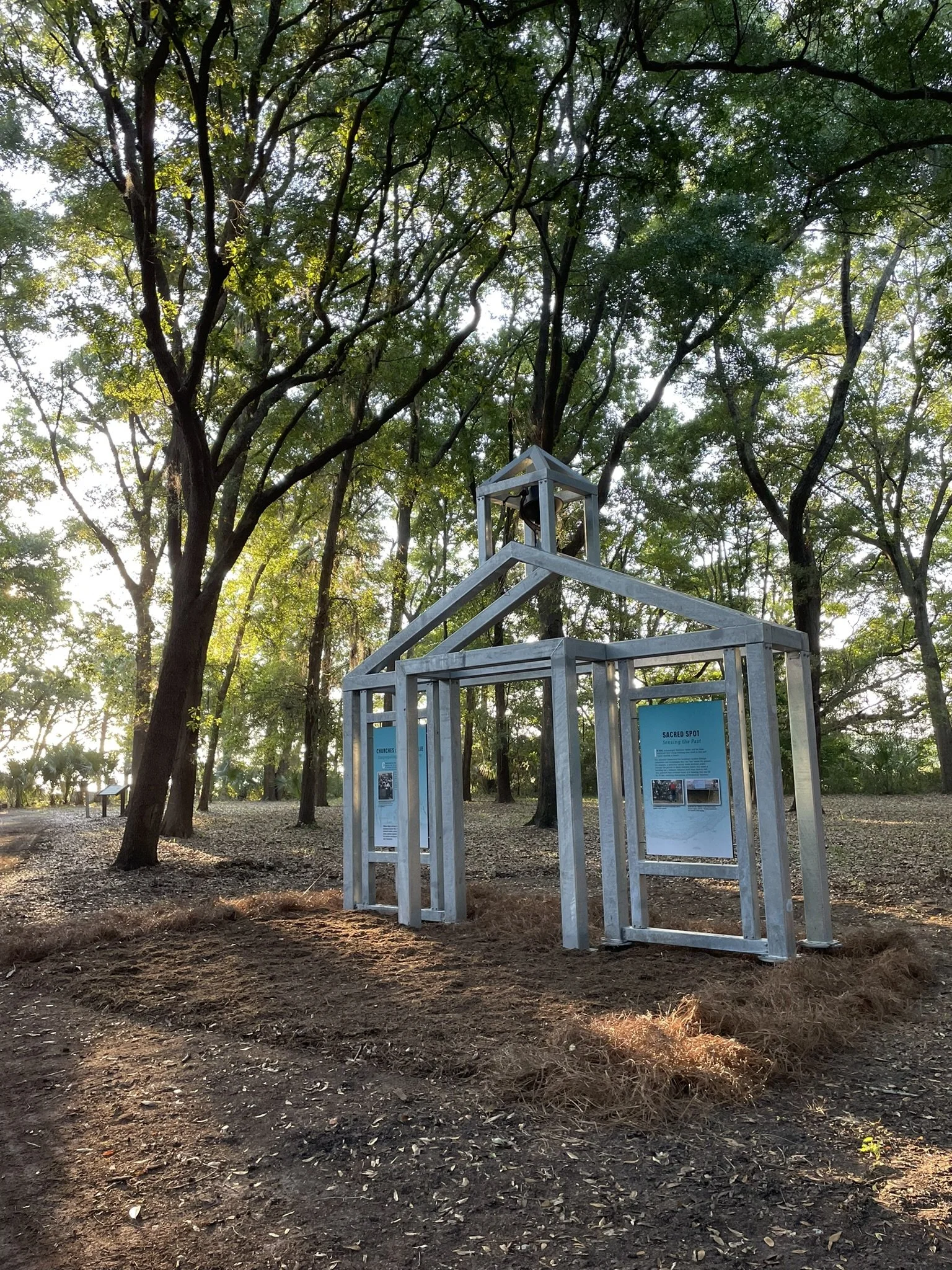 A metal outline of the front of a traditional small church with exhibit panels where windows would be, and a church bell hanging from the steeple. Sun is streaming through big live oak trees in the background. 