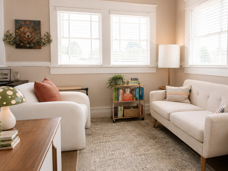 A cozy living room featuring two white sofas with colorful throw pillows, a wooden side table with a green and white polka-dot lamp, a small bookshelf with books and decorative items, a tall white floor lamp, and a framed bear portrait on the wall.