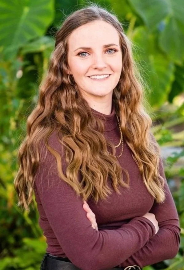 A young woman with long, wavy brown hair wearing a purple long-sleeve top, standing outdoors with green foliage in the background, crossing her arms and smiling.