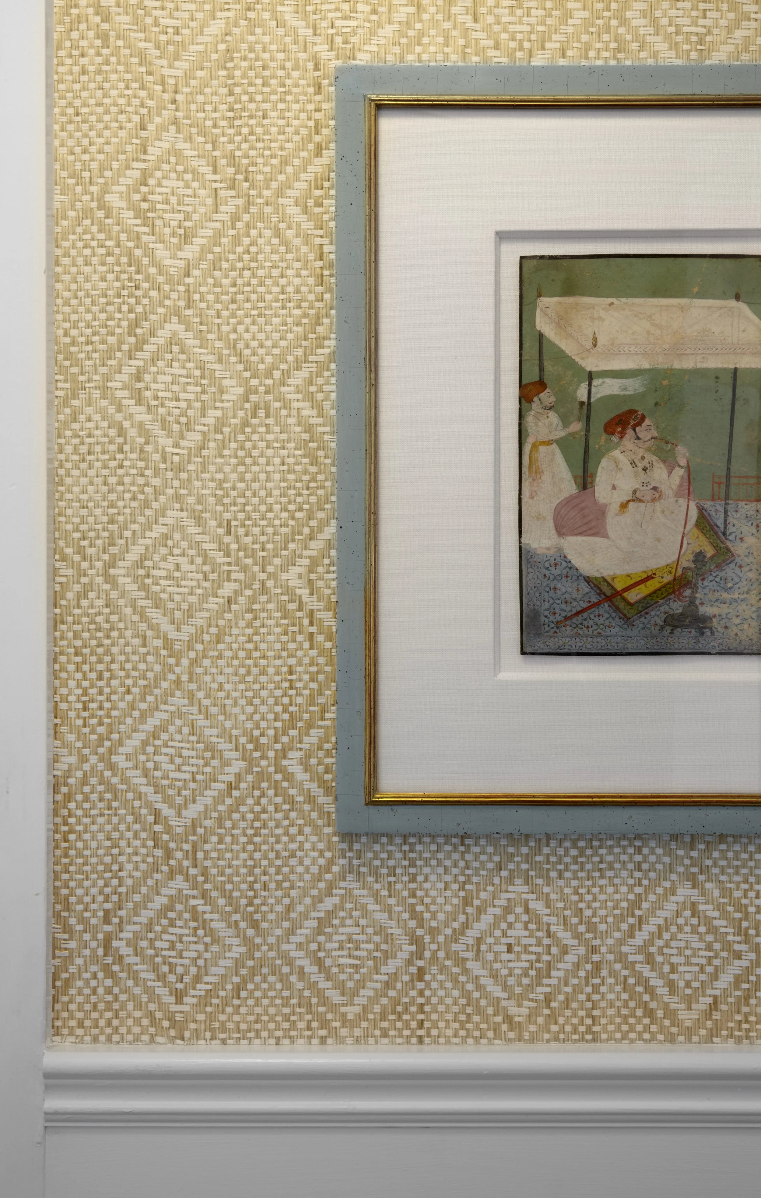 Close-up of a framed artwork on a wall with beige woven diamond-patterned wallpaper. The artwork depicts a scene with two figures, one seated and playing a musical instrument, and a standing figure in the background.
